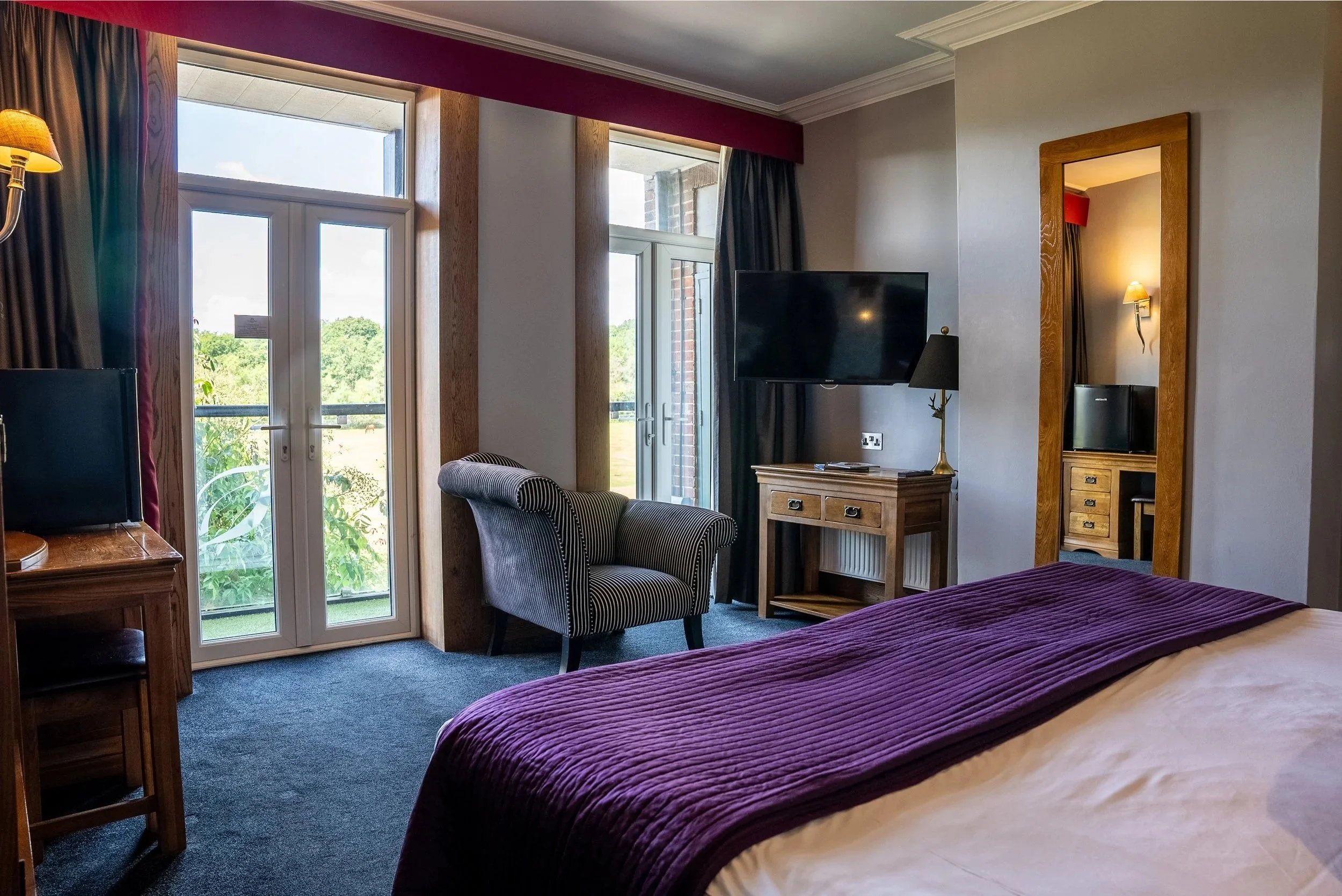 Interior of a hotel room with a bed, a striped armchair, a wooden bedside table, a wall-mounted television, a standing lamp, a large mirror, and glass doors leading to a balcony with a view of green trees and a cloudy sky.