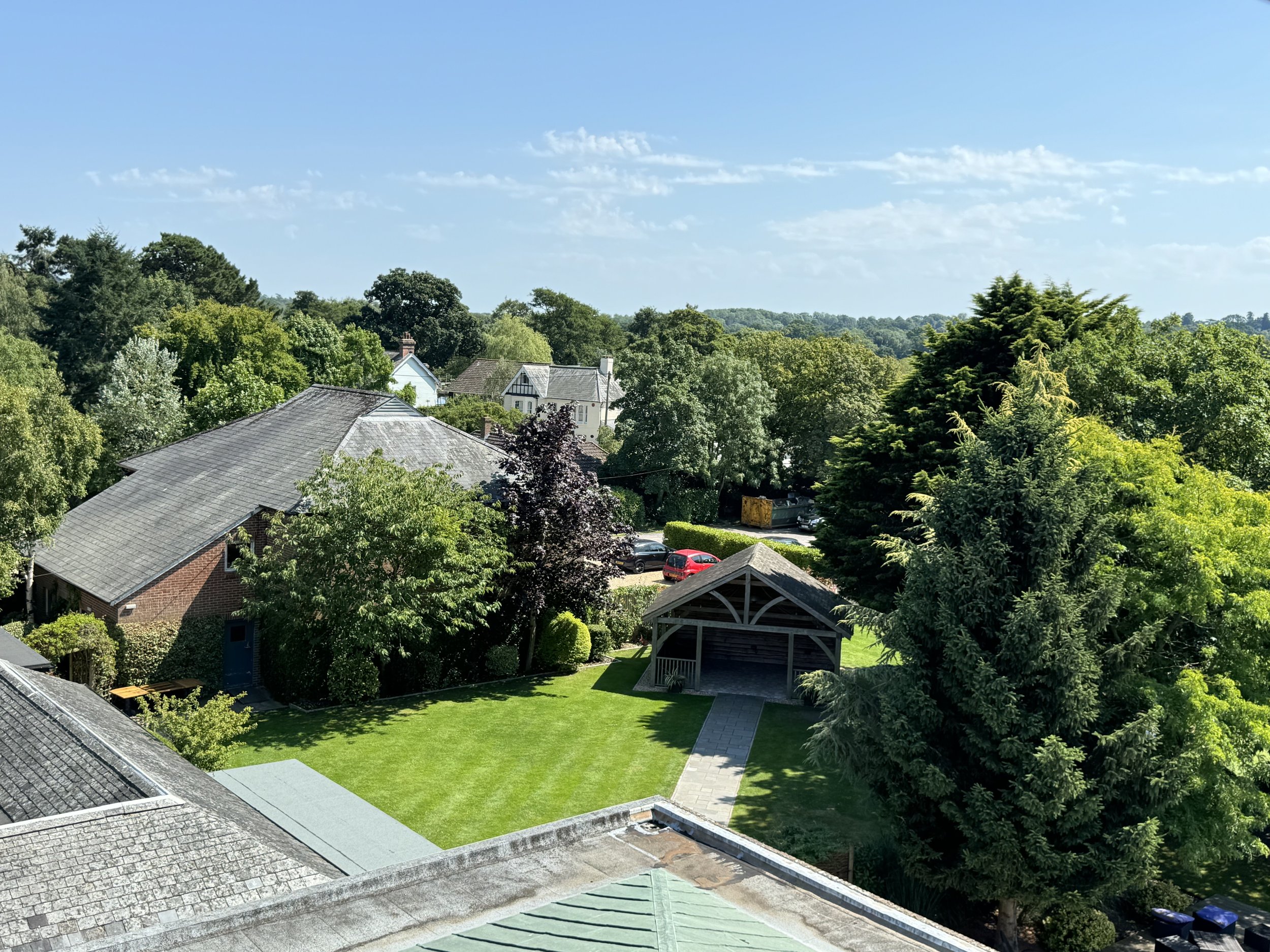 Aerial view of a lush green residential area with several houses, trees, a well-maintained lawn, a small wooden shed, and parked cars under a clear blue sky.