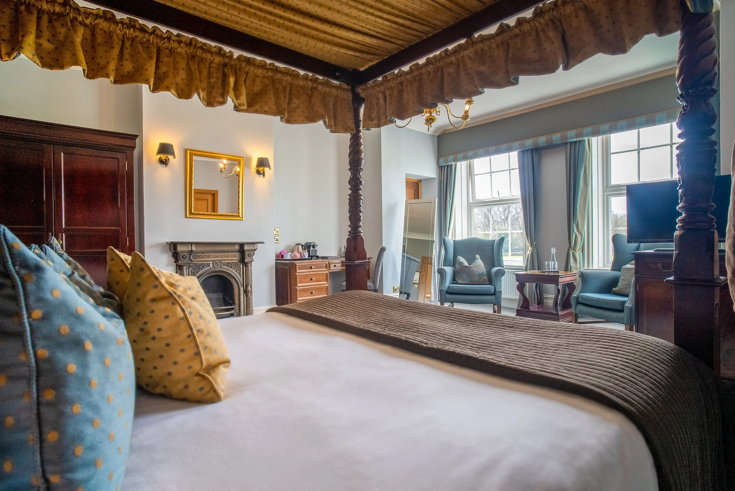 A bedroom with a four-poster bed, decorated with blue and yellow polka dot pillows, a brown bed cover, and a white sheet. The room has a fireplace, a mirror, a small wooden table, chairs, a TV, and large windows with curtains, allowing natural light 