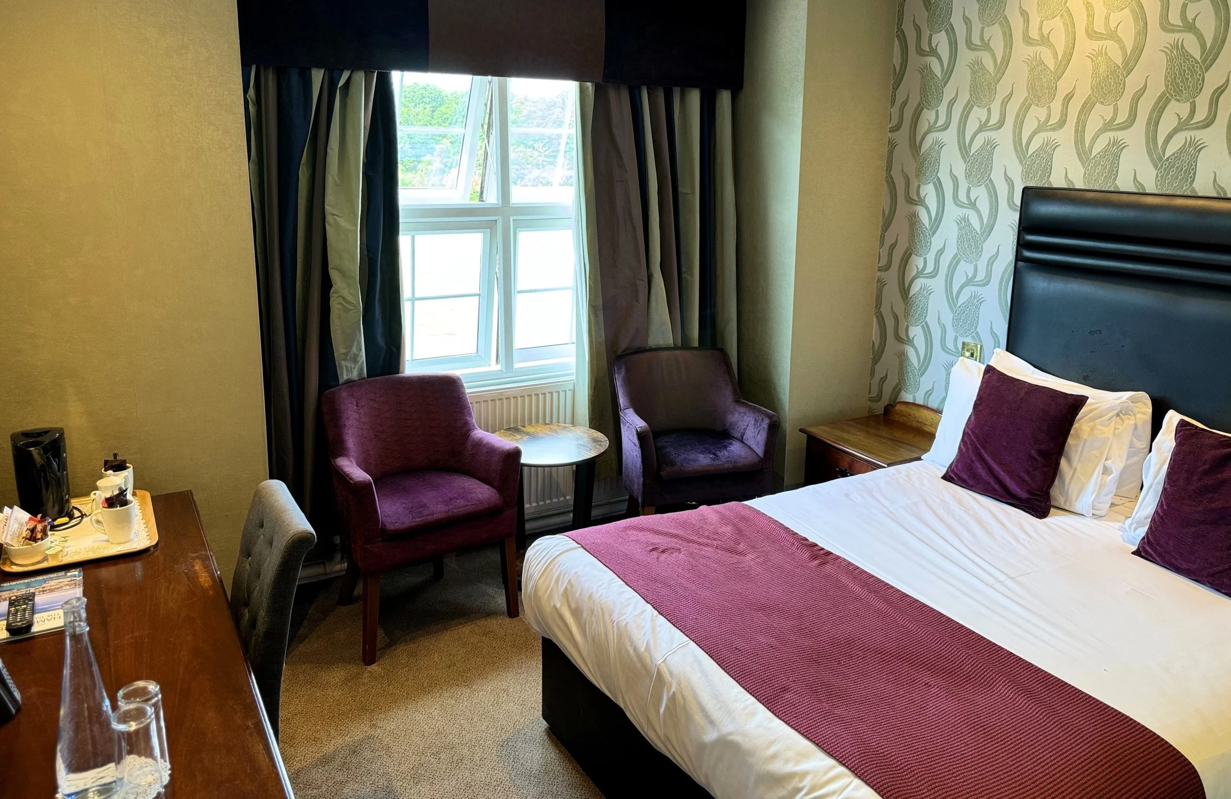 Hotel room with a bed, purple pillows, a window with curtains, two purple armchairs, a small side table, a wooden desk with tea and coffee supplies, and patterned wallpaper.