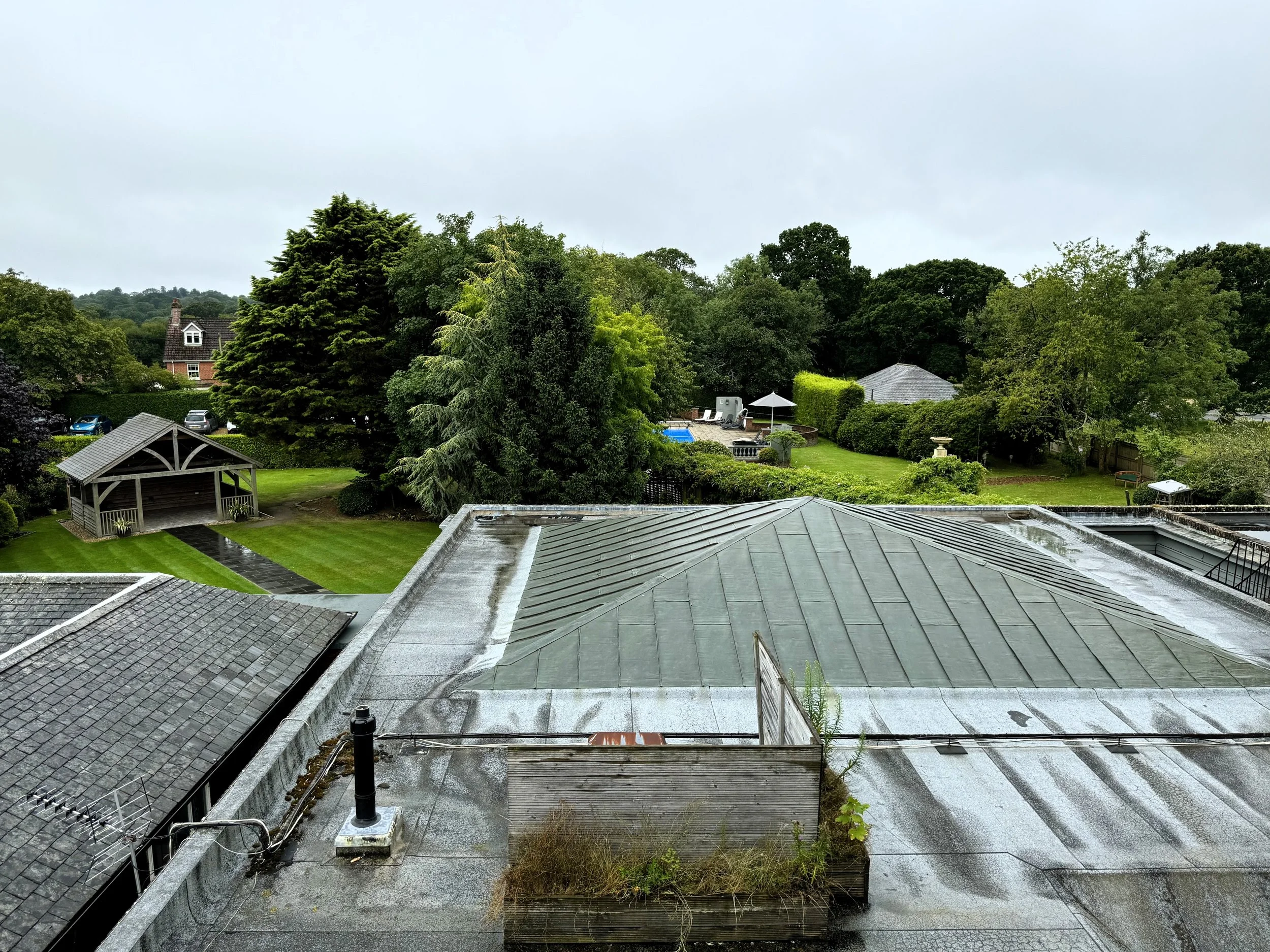 View from a rooftop showing a rain-soaked metallic roof, a lush green backyard with trees, a gazebo, and outdoor furniture, under a cloudy sky.