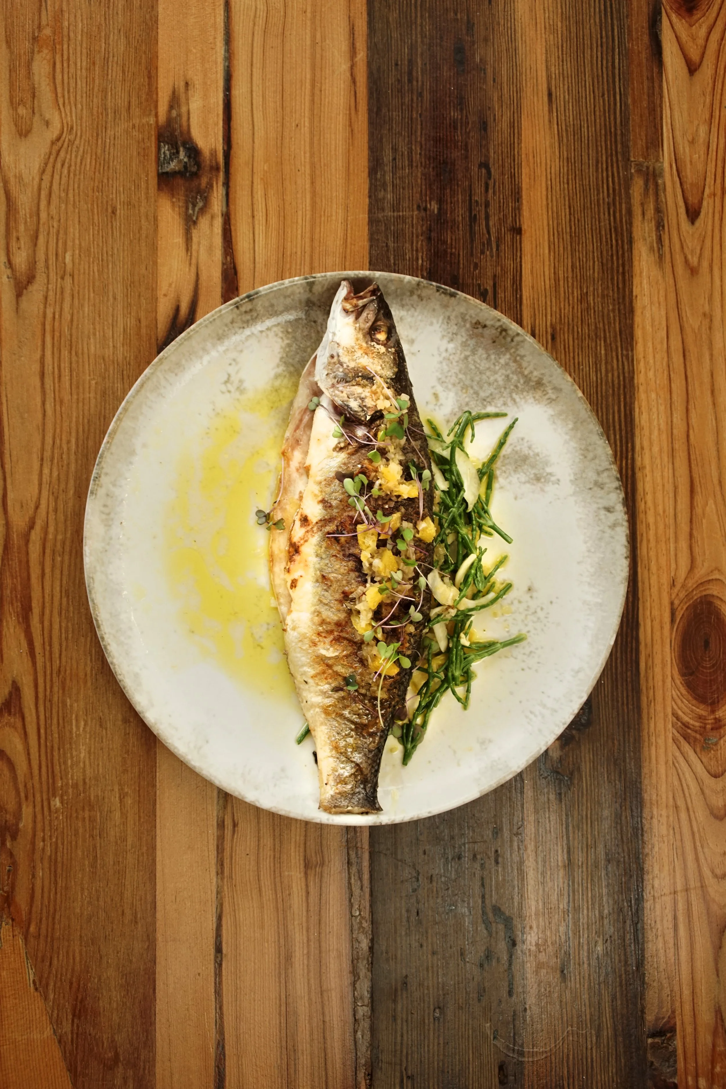 A cooked whole fish garnished with herbs and lemon slices, served on a white plate with a drizzle of yellow sauce, placed on a wooden table.