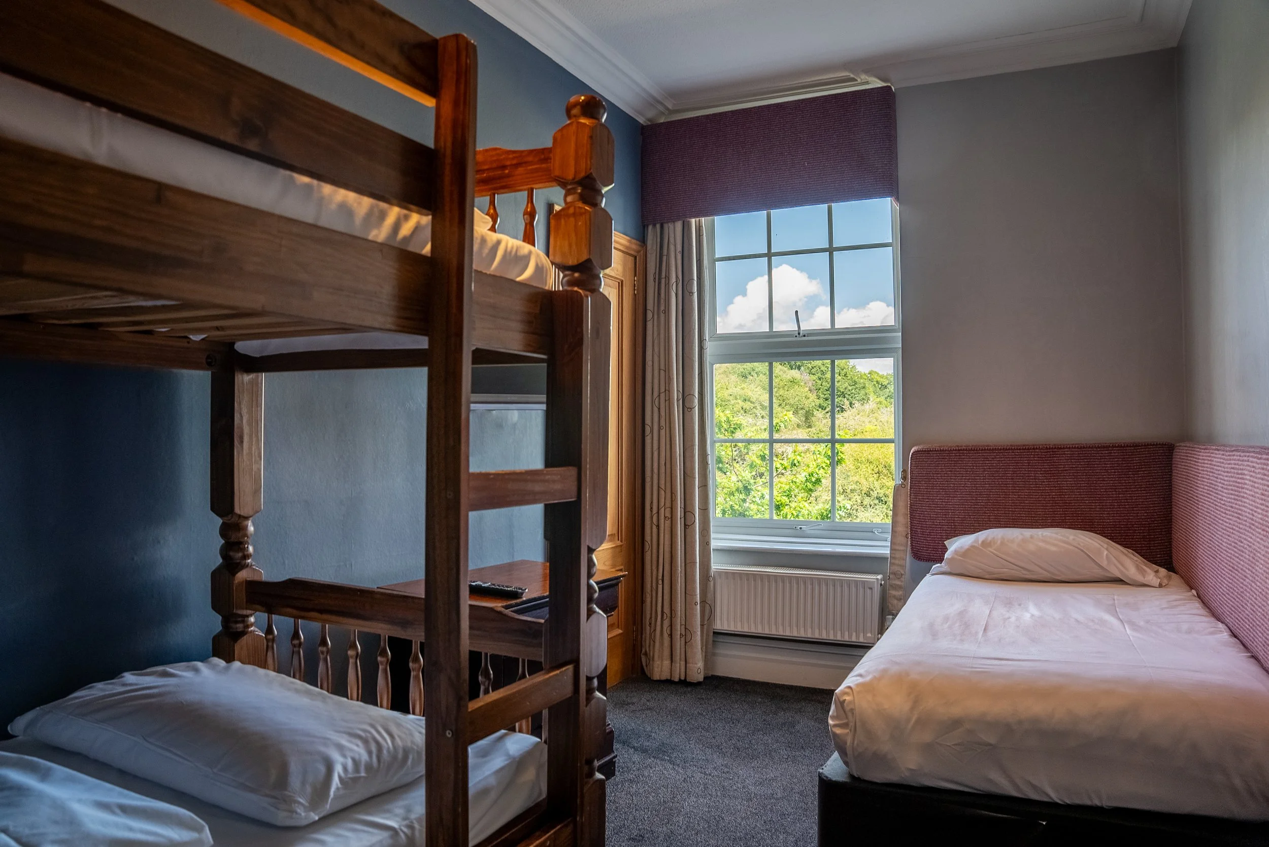 A bedroom with a bunk bed on the left, a single bed on the right, and a large window showing green trees outside.