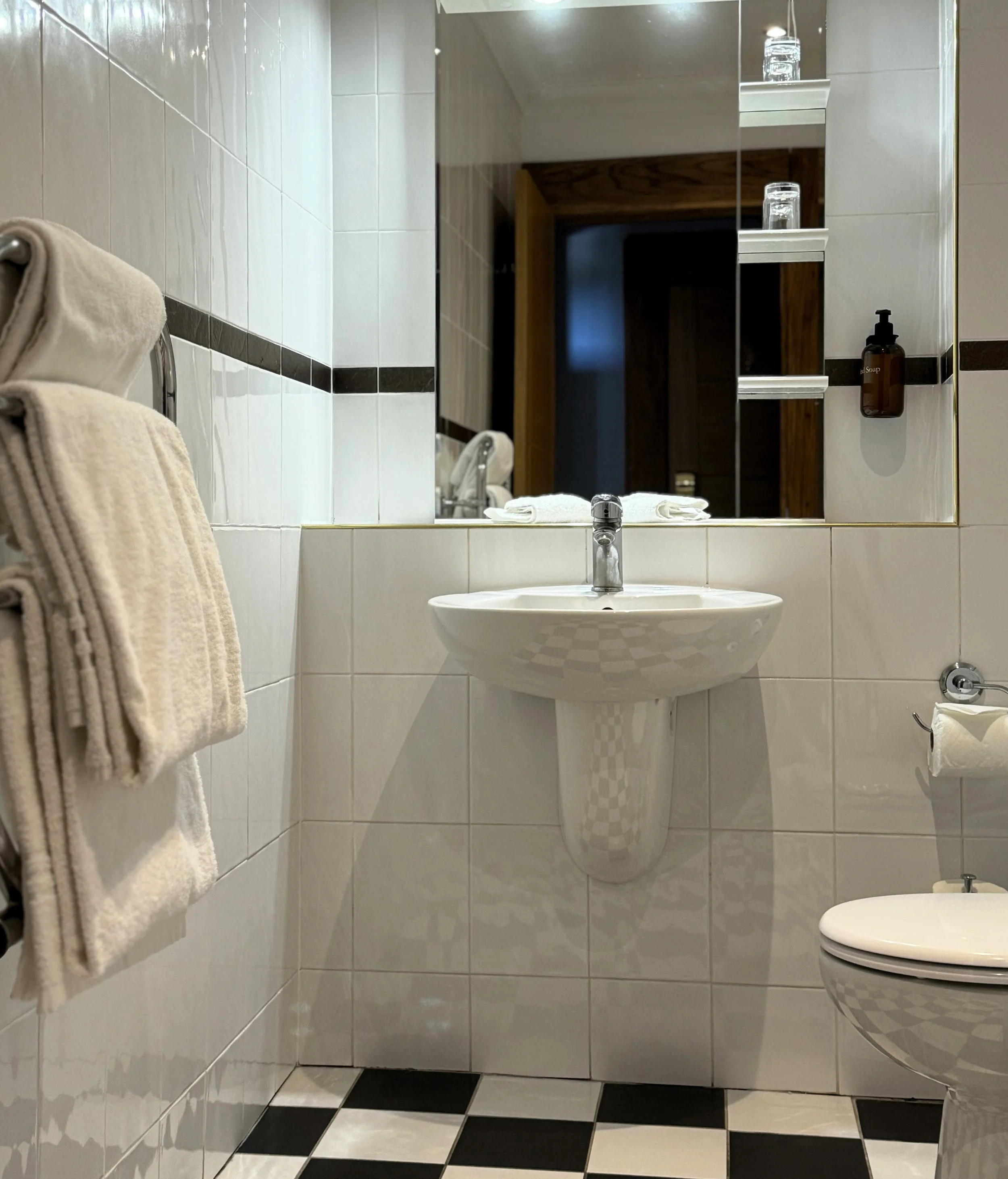 A small bathroom with a white tiled wall, a mirror, a sink, and a toilet. Towels are hanging on a rack on the left, and shelves with toiletries are reflected in the mirror. The floor has a black-and-white checkered pattern.