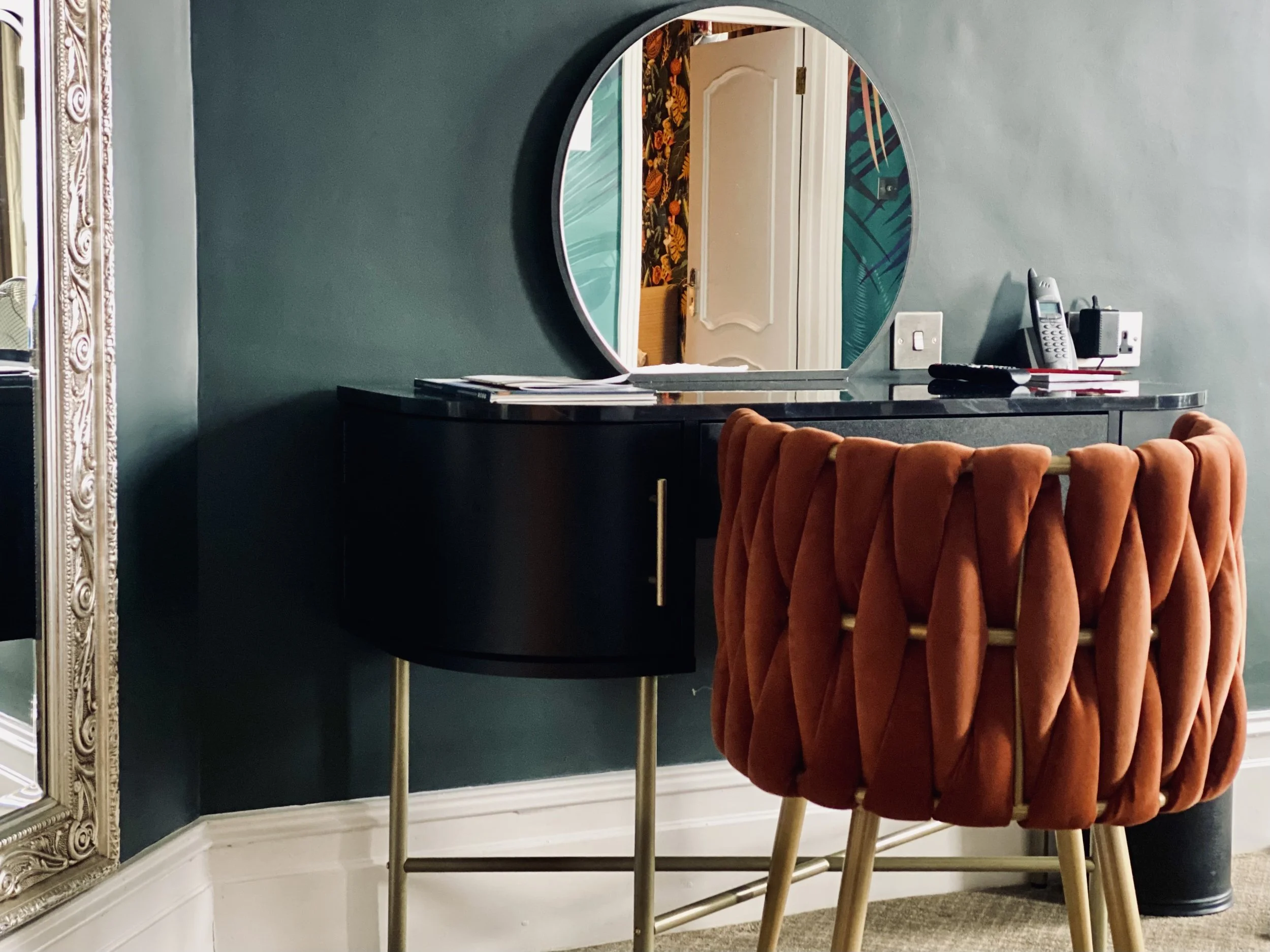 A black vanity table with a small gold handle, positioned against a green wall, with a round mirror above it. On the table, there are some papers, a cordless phone, and electronic devices. An orange, plush, tufted chair with gold legs is in front of 