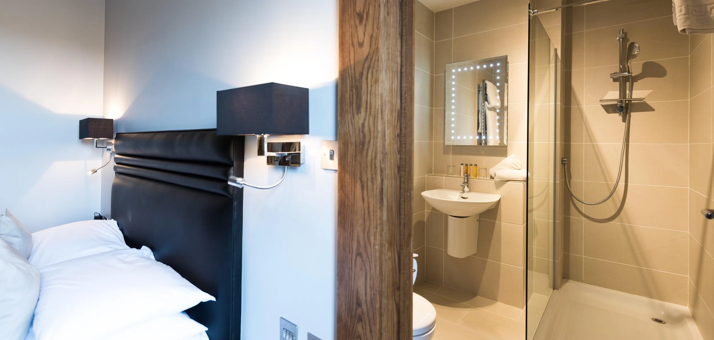 A hotel room with a bed on the left and an attached bathroom on the right, including a shower, sink, and mirror with LED lights.