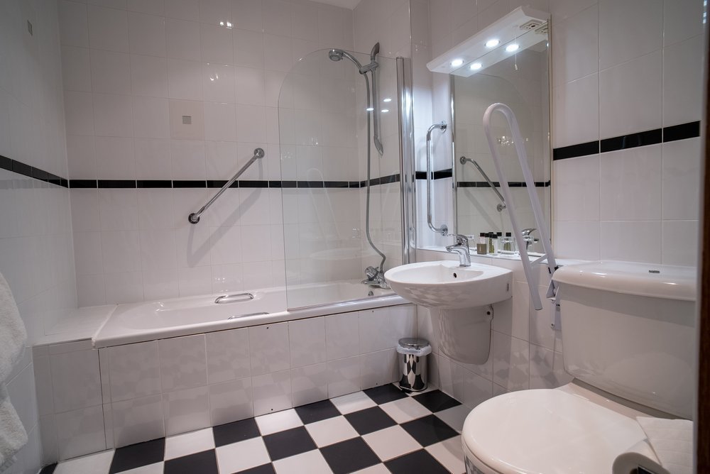Accessible bathroom with a bathtub, grab bars, a sink with a mirror, and a toilet, featuring black and white checkered floor tiles.