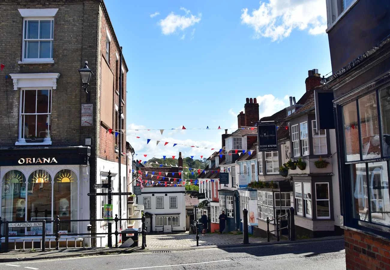 A street scene with multicolored triangular bunting hanging above, surrounded by buildings with storefronts and houses, and a clear blue sky with clouds.