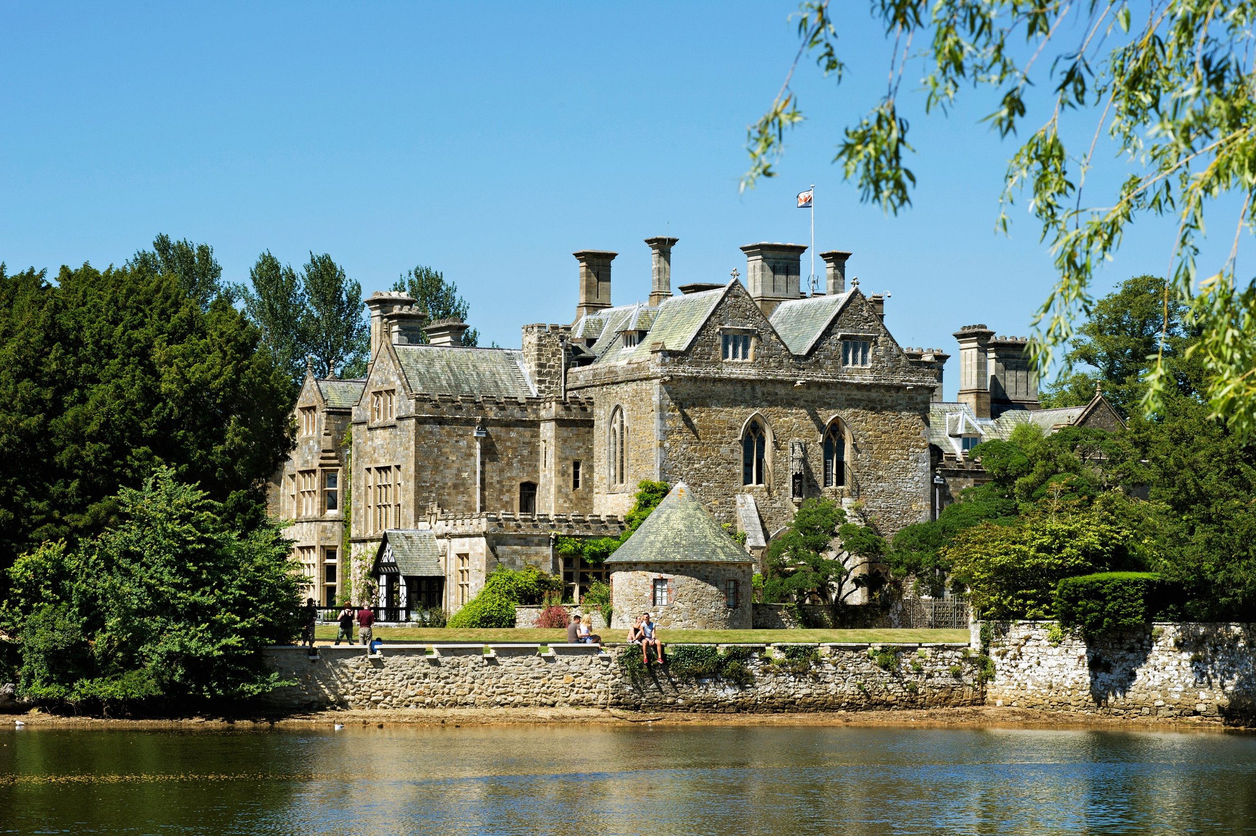 A historic stone castle with multiple towers and chimneys, surrounded by trees and situated by a body of water under a clear blue sky.
