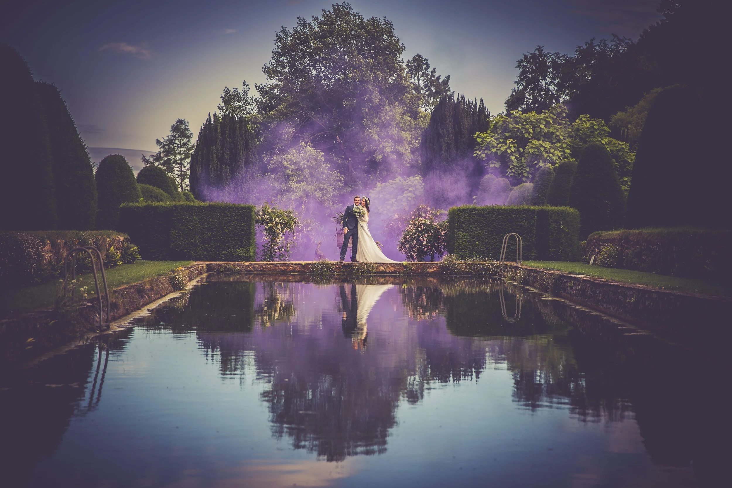 Couple in wedding attire standing by a reflection pool in a lush garden with trees and purple smoke in the background.
