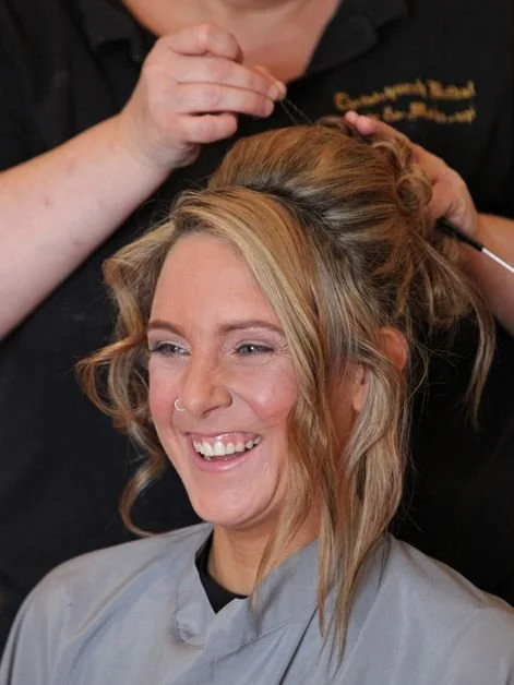 A woman with blonde wavy hair smiling while getting her hair styled at a salon.