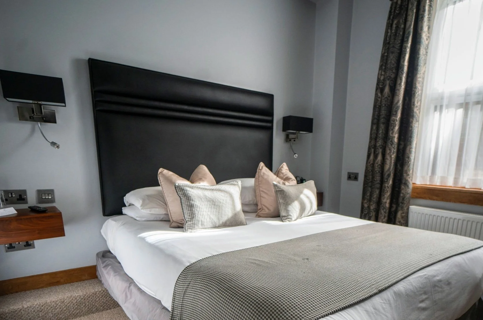 A neatly made bed with multiple pillows in a hotel room, black headboard, nightstands with lamps and a window with curtains.