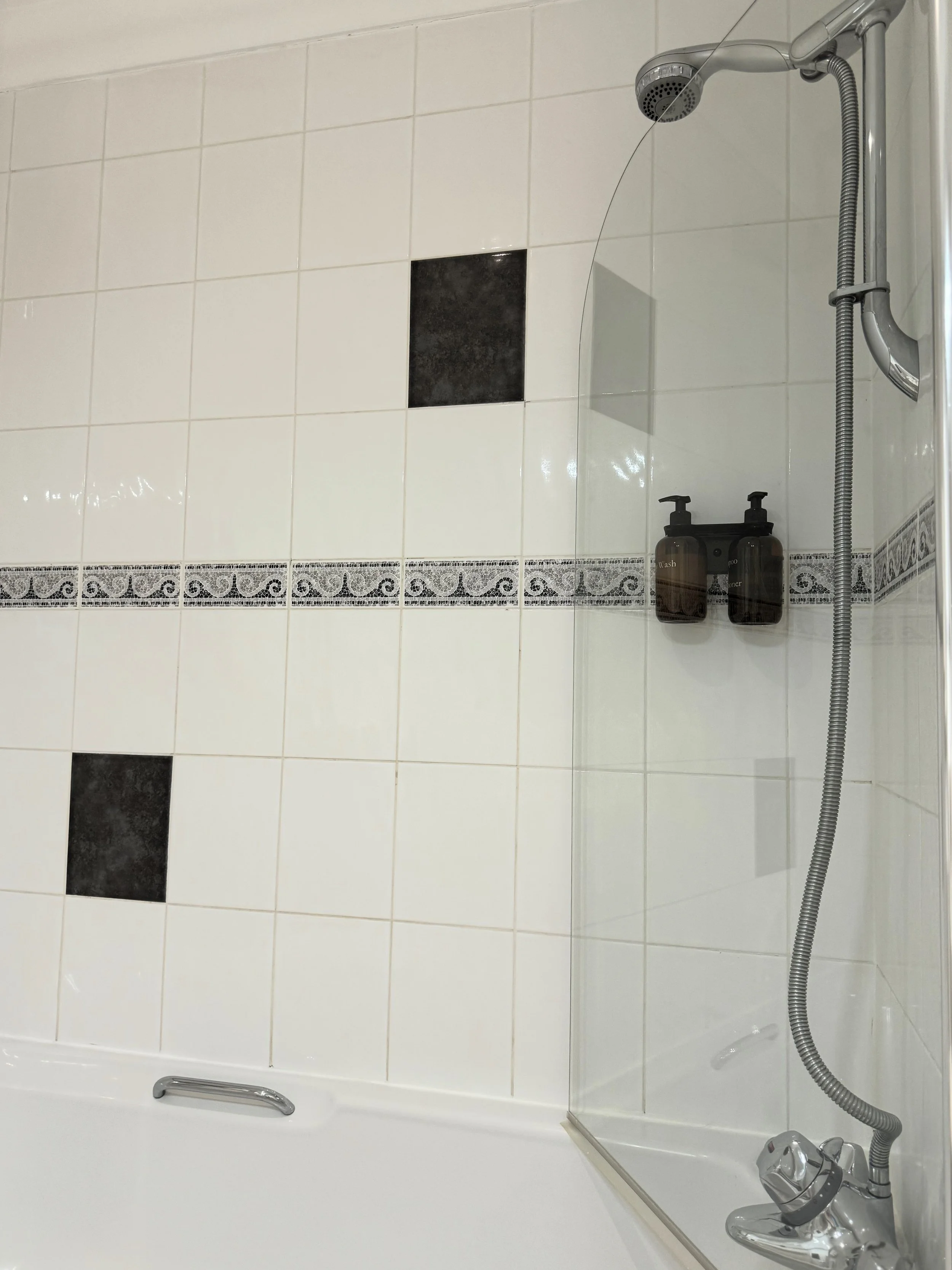 Shower area with white tiled wall, black accent tiles, glass partition, and wall-mounted soap dispensers.