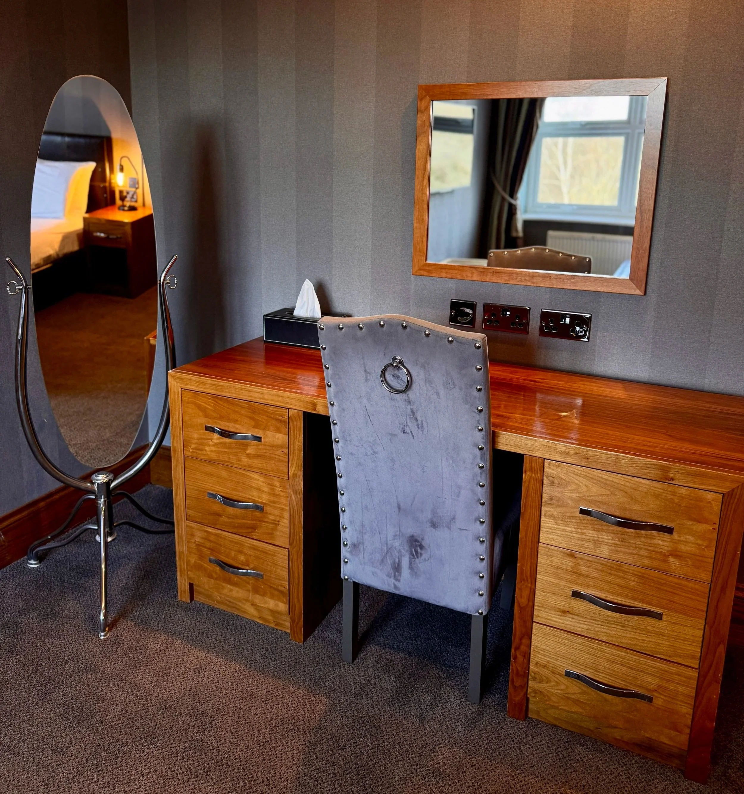 A bedroom with a wooden desk, a grey upholstered chair, a tall wooden wardrobe, a wall-mounted mirror, and a standing mirror.