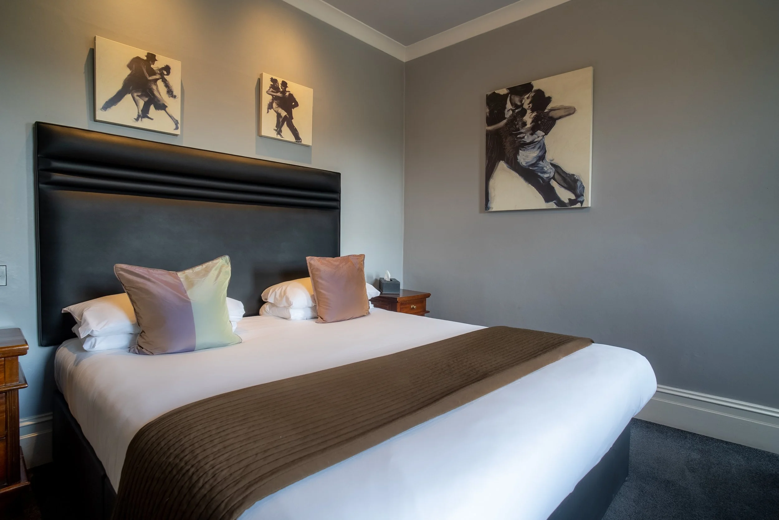 A neatly made hotel bed with white linens, a brown bed runner, and two beige pillows, against a gray wall. Above the bed are three black and white paintings, with the large one depicting two dancers.
