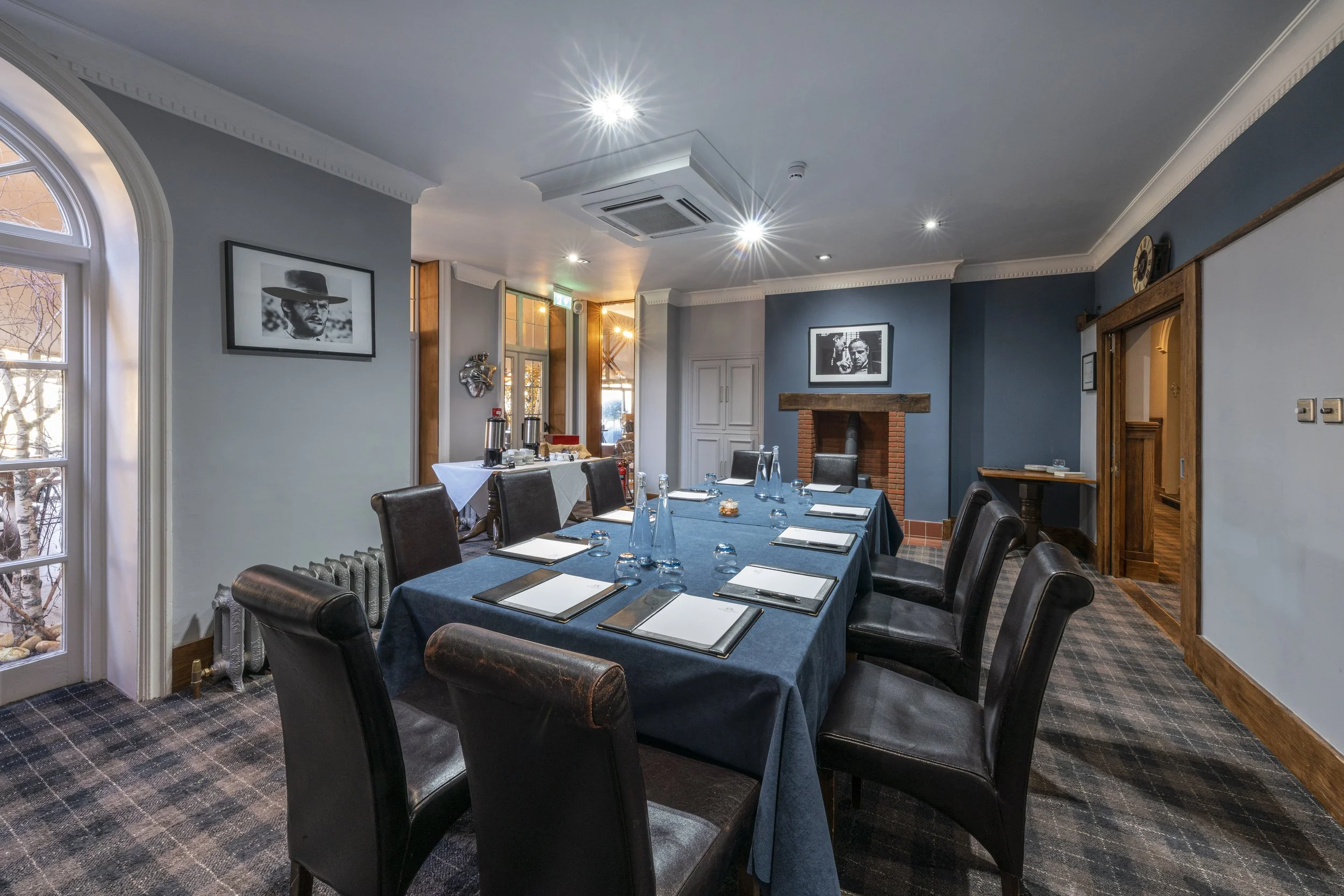 A conference room with a long table covered with a dark blue cloth, set with glasses and notepads, surrounded by leather chairs, large window with an arch, artwork on the walls, and a brick fireplace.
