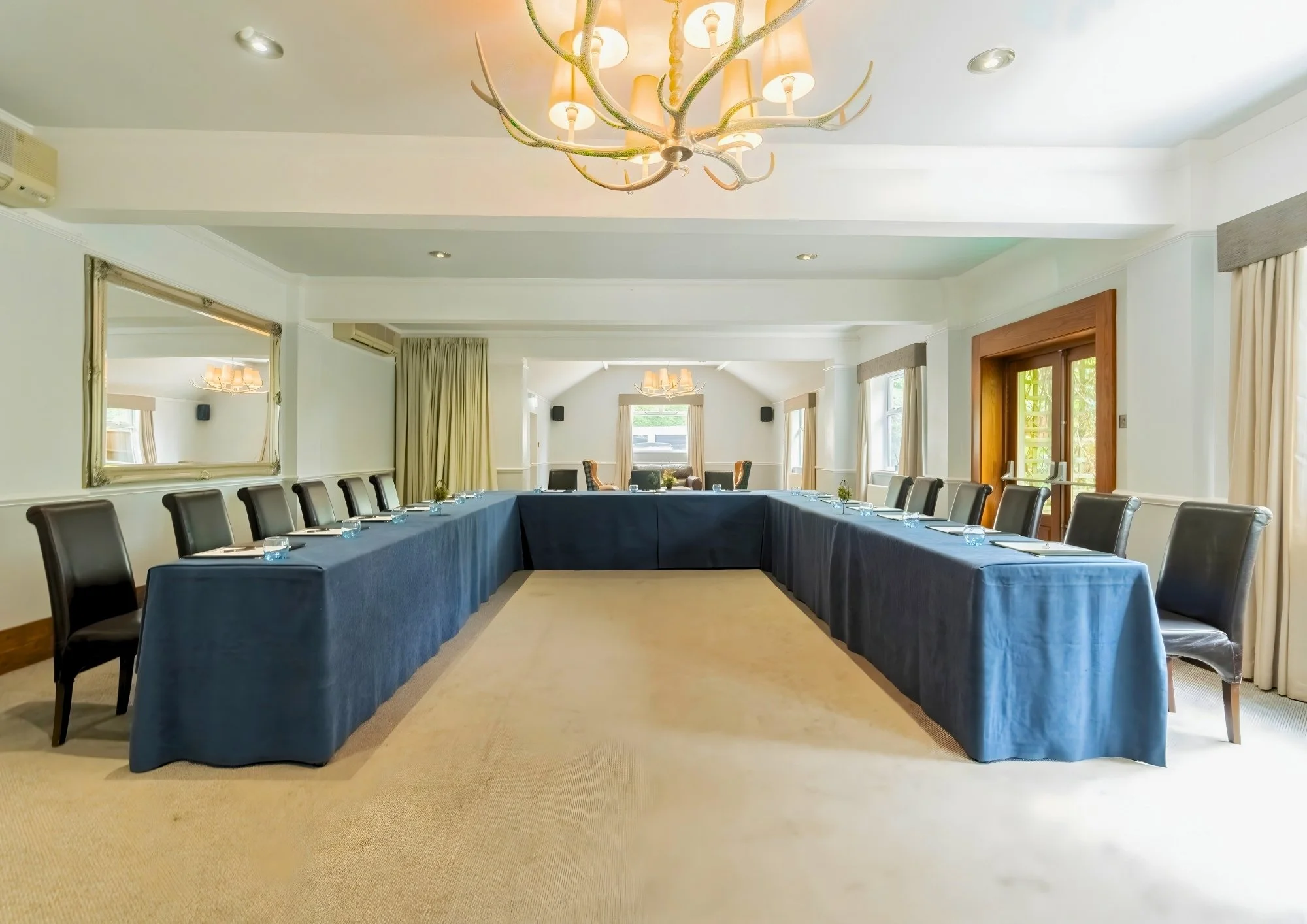 Elegant conference room set up with long tables covered in dark blue tablecloths, chairs with black covers, a chandelier, two large screens, a mirror, and a door, with a small floral arrangement on the front table.