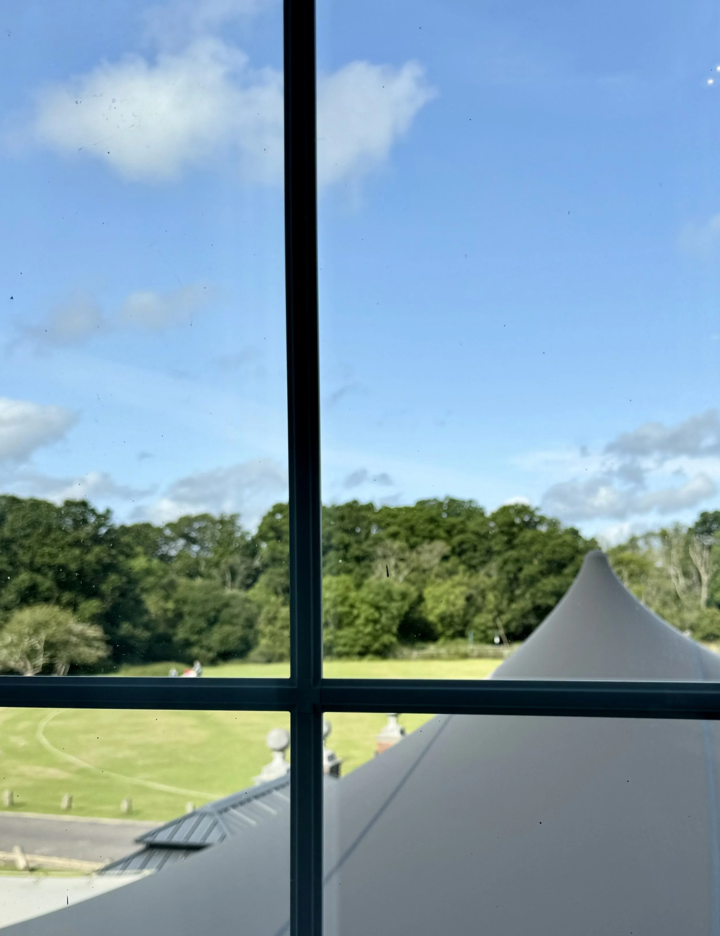View through a window showing a green park with trees, a path, and a tent, with a blue sky and clouds above.