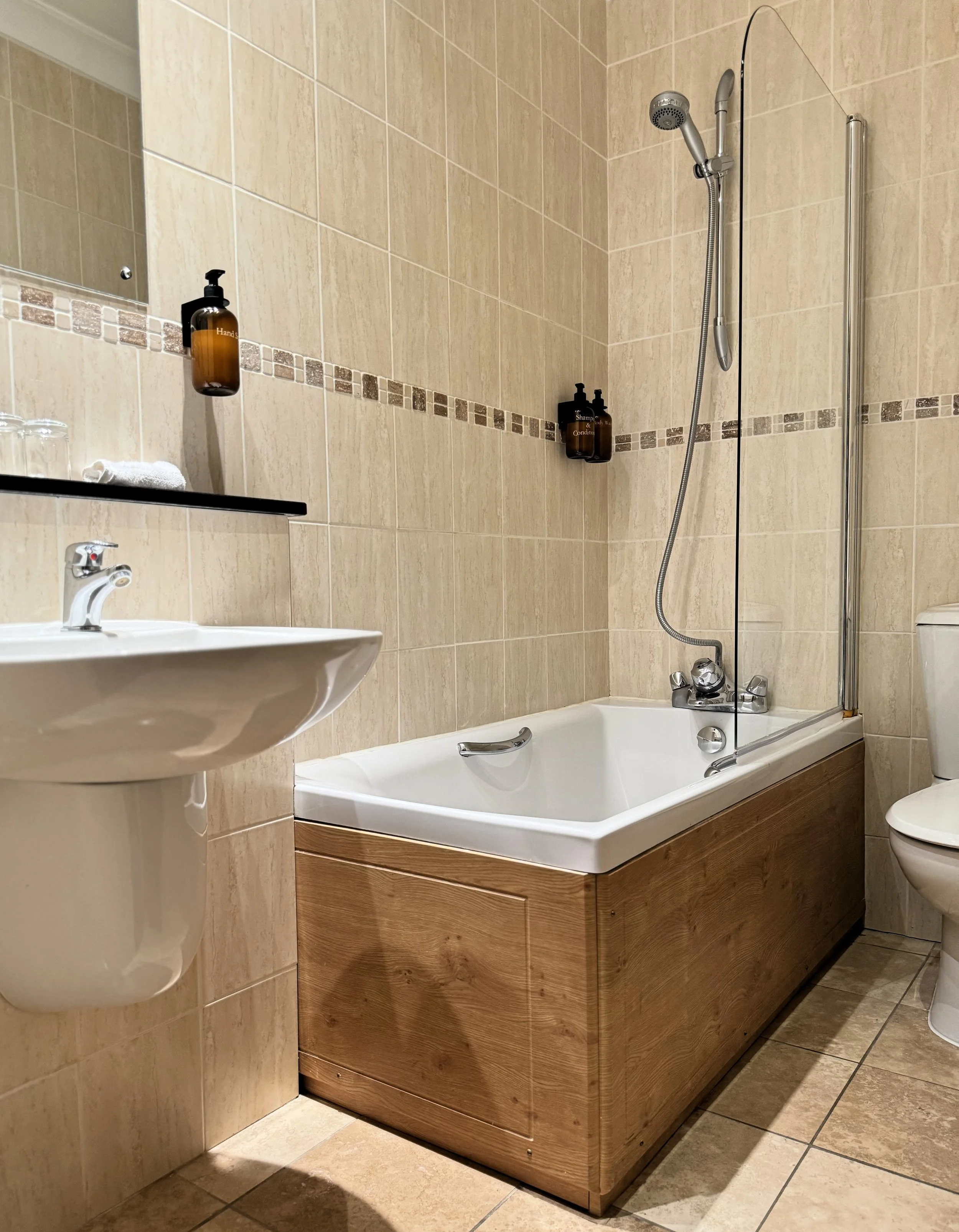 Bathroom with a bathtub, shower head, wooden panel on the side of the tub, white sink, mirror, and beige tiled walls and floor.