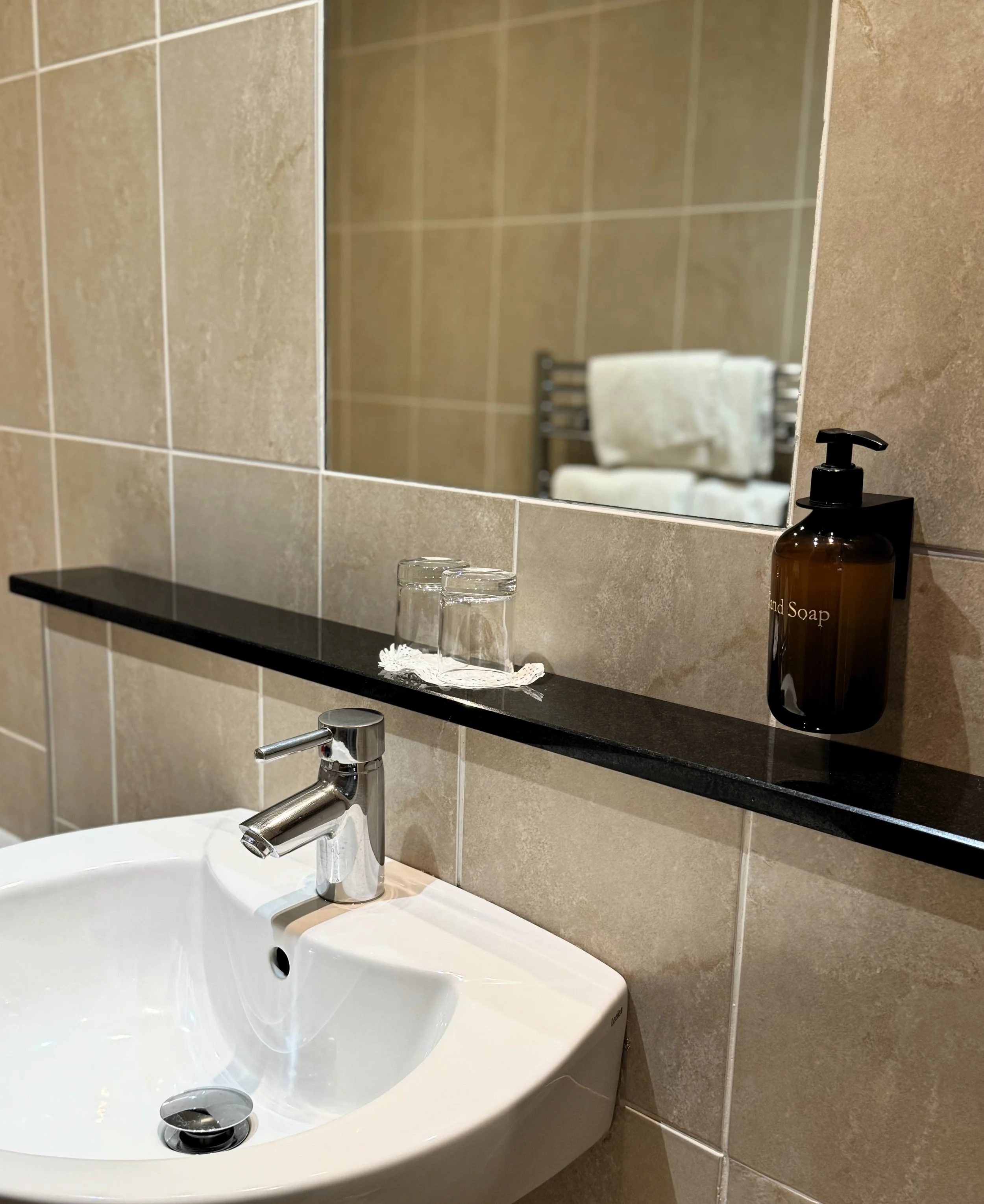 Bathroom sink with a chrome faucet, a black shelf above holding two glasses on a lace doily, and a brown soap dispenser labeled 'Hand Soap'; a large mirror reflecting a towel rack with white towels.