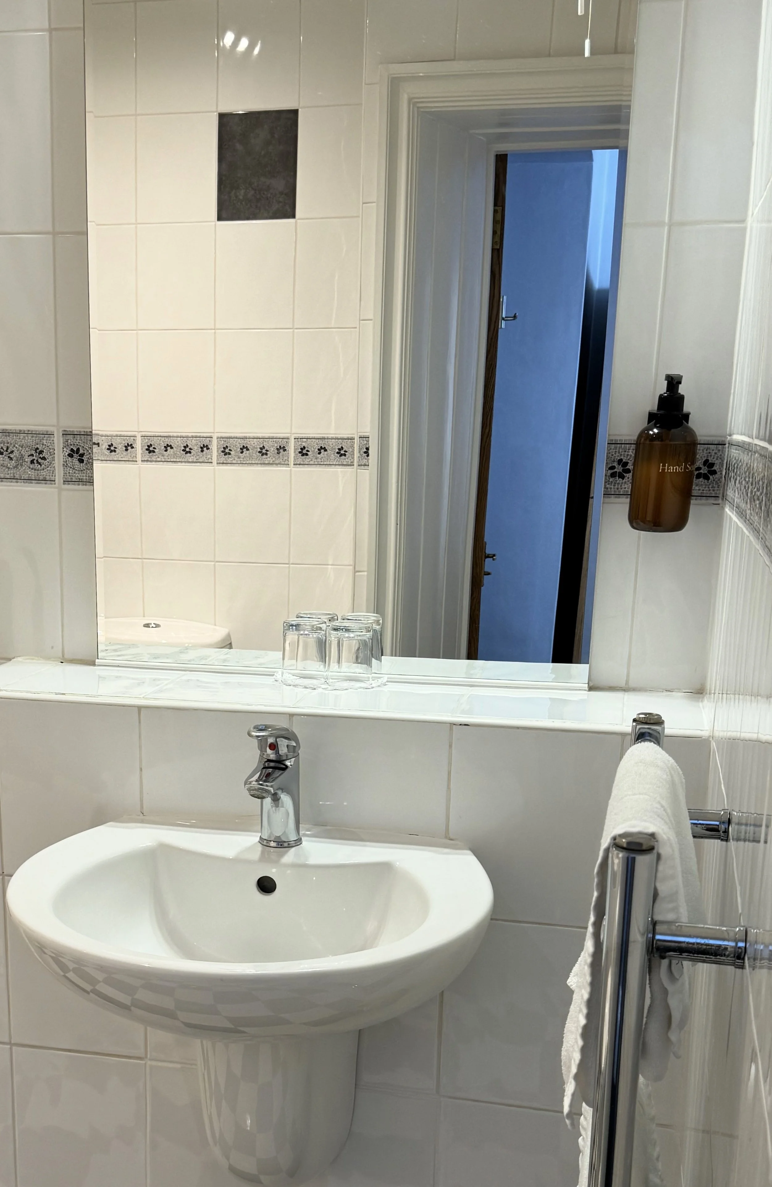 A bathroom with a white sink, a mirror, and a hand soap dispenser on the wall. There are three clear glasses on the sink's counter and a towel hanging on a rack. The reflection in the mirror shows a partially open door leading to another room.