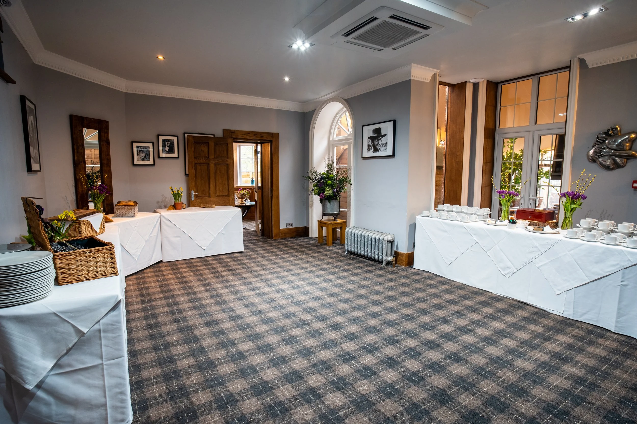 A decorated dining room with a long table covered with a dark cloth, set with notebooks, pens, and glassware, and adorned with multiple floral centerpieces. There are black leather chairs around the table. To the left, a large flat-screen TV displays an image of two people sitting on a staircase. The room has white walls, wood paneling, large windows with white frames, and a painting of an archway hallway on the wall.