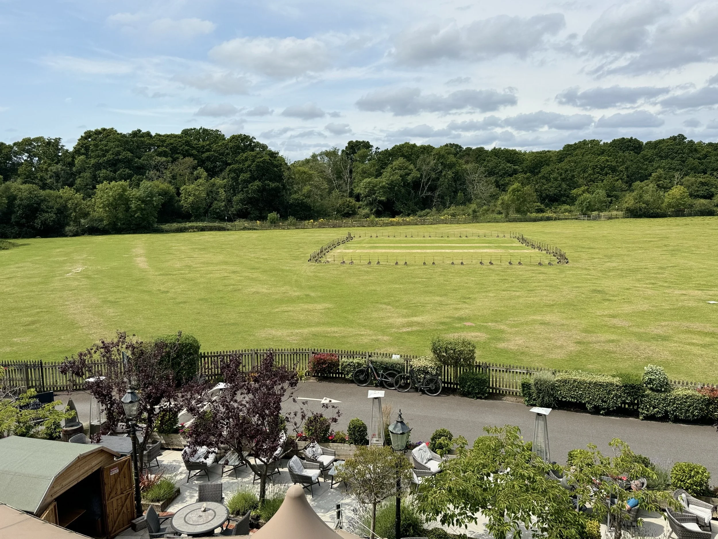 A view of a large grassy field with some fencing and trees in the background, and a patio area with chairs, tables, and potted plants in the foreground.