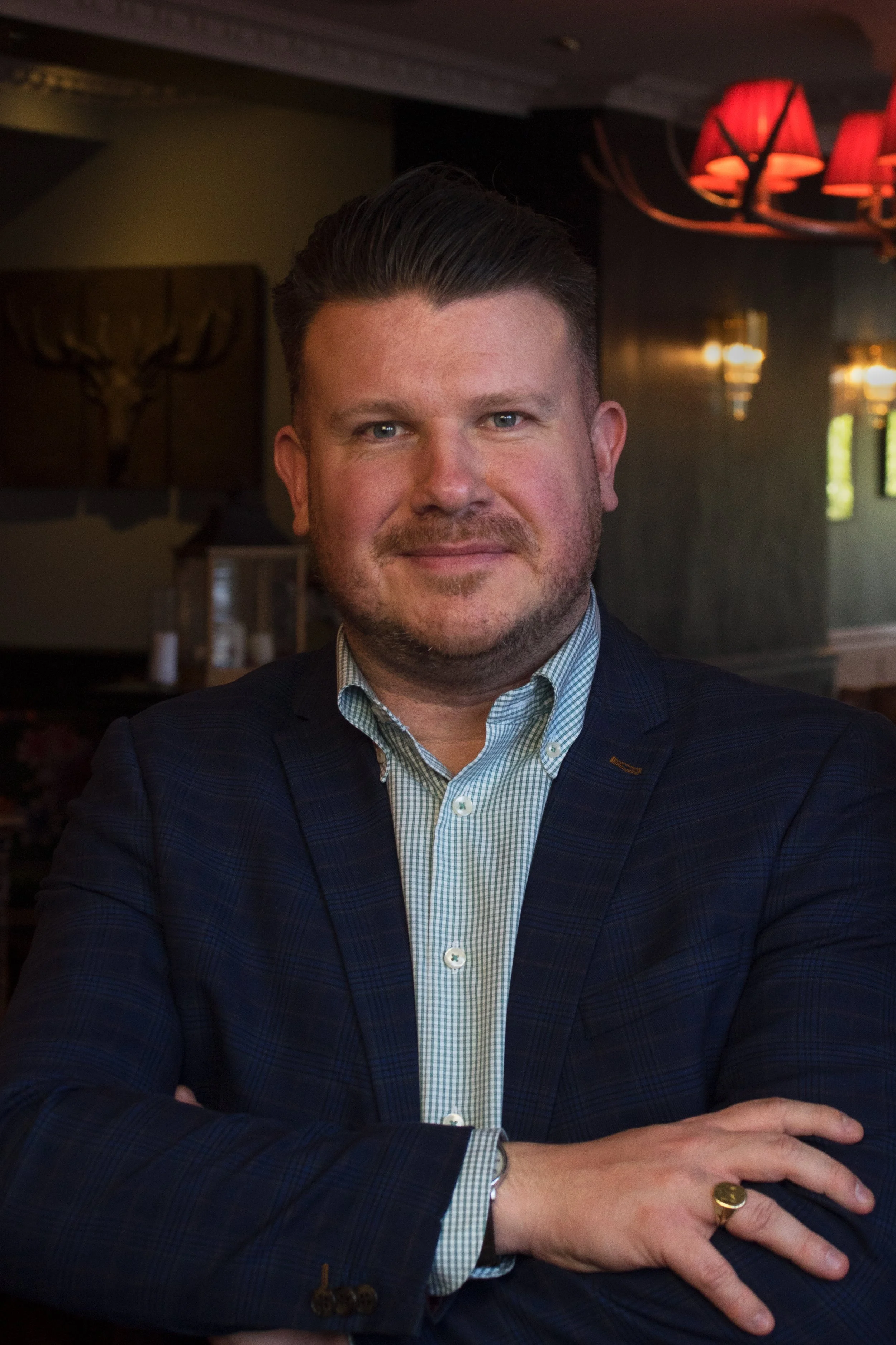 A man with short brown hair and a beard posing outdoors in front of trees, wearing a dark blazer, light blue checkered shirt, and a pocket square, with arms crossed.