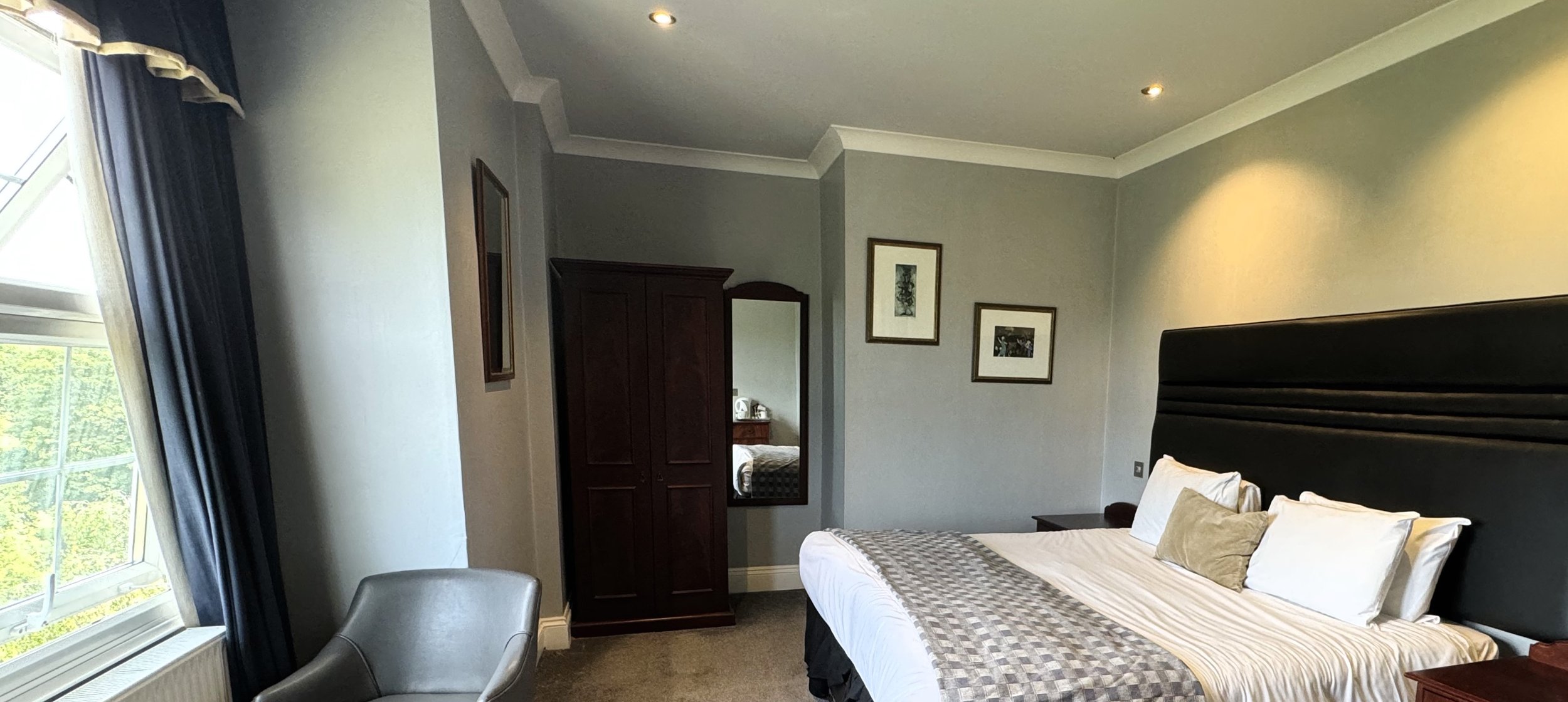 A neatly made bed with white sheets and multiple pillows in a cozy hotel room. A dark headboard is against the wall, with a small nightstand beside it. The room has framed pictures on the grey walls, a wardrobe, and a mirror. A window with blue curta