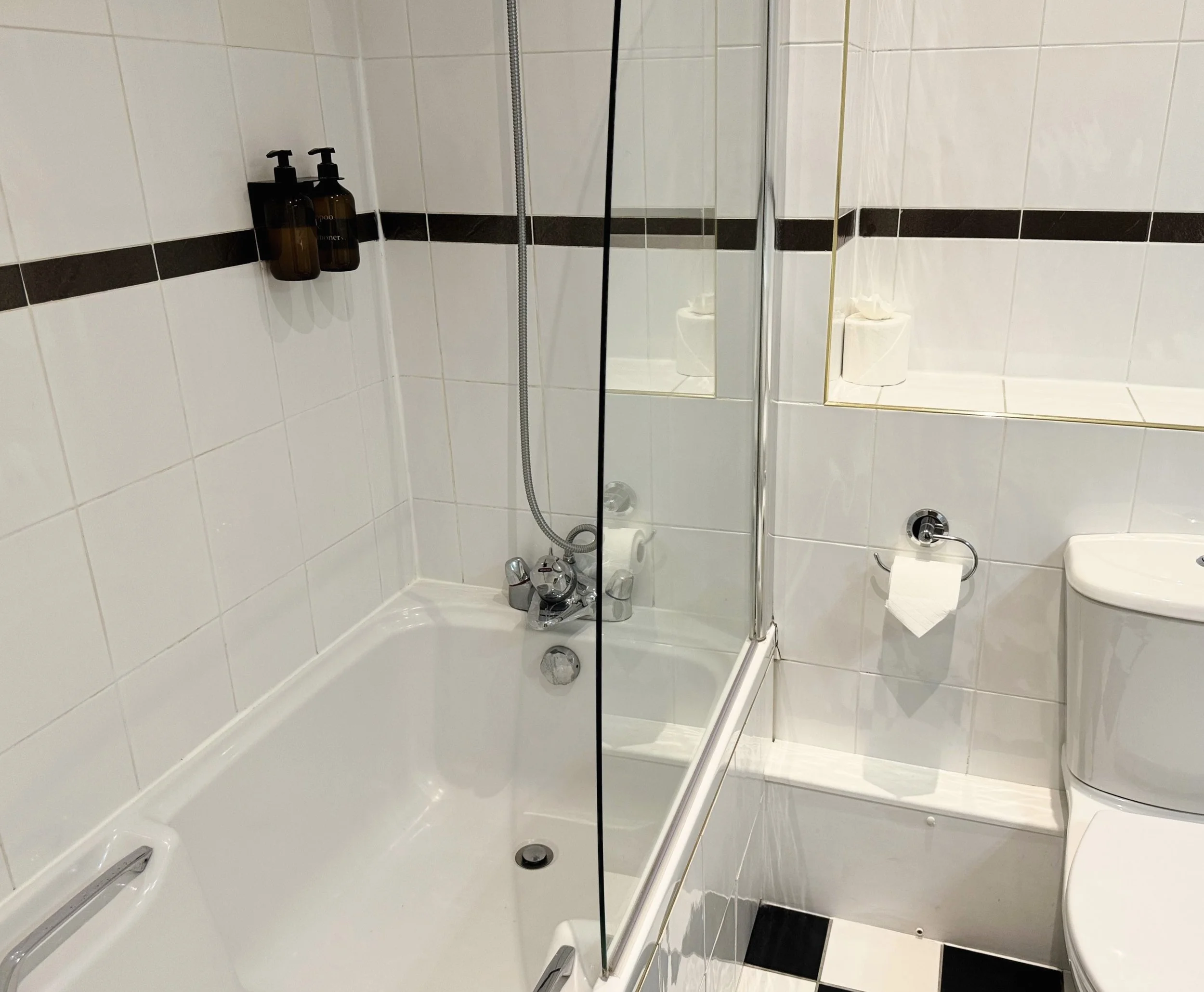 A bathroom with a bathtub on the left, a showerhead with a flexible hose, a mirrored wall, and a toilet on the right. The walls are tiled with white tiles accented by a dark horizontal stripe. There are two brown bottles on a wall-mounted shelf in th