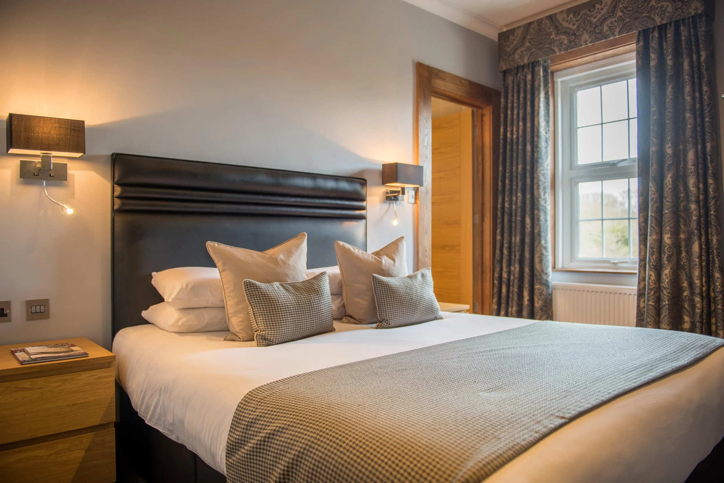 A neatly made bed with multiple pillows in a cozy hotel room, illuminated by wall-mounted lamps, next to a window with patterned curtains.