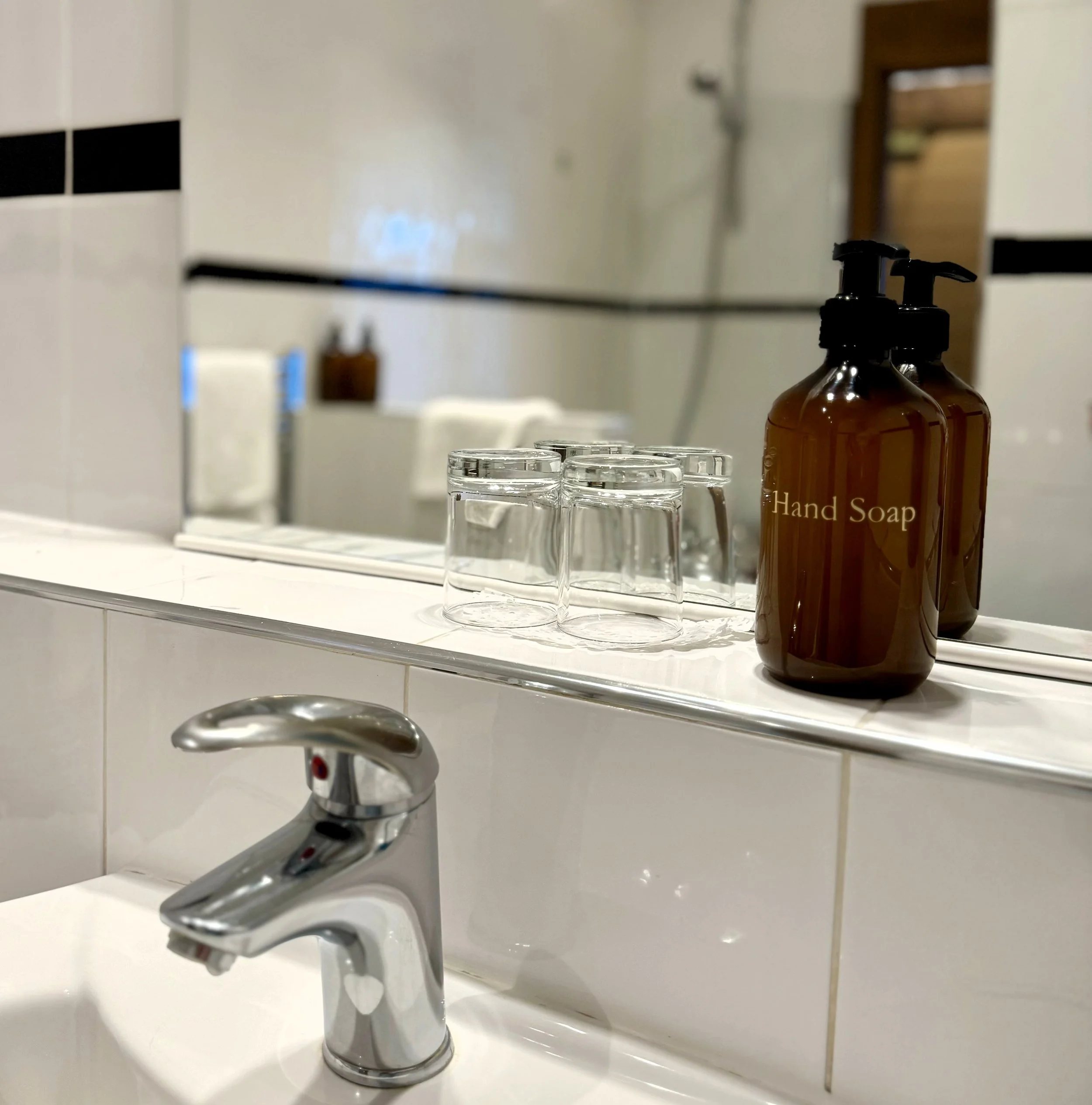 Bathroom sink with a silver faucet, a brown bottle labeled Hand Soap, and three upside-down glasses on a white tiled counter, with a mirror and shower in the background.