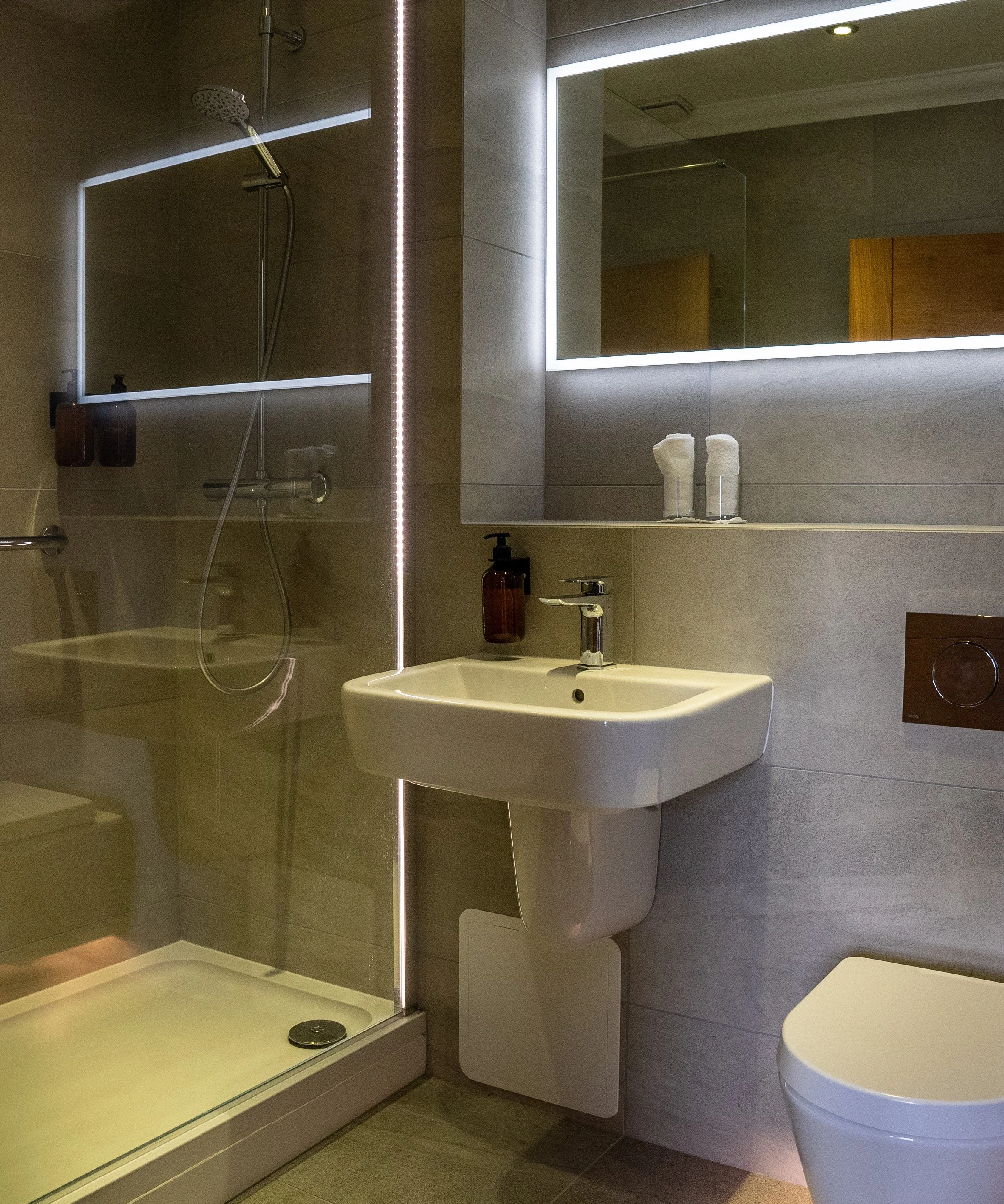 A modern bathroom with a glass-enclosed shower area, a ceramic sink with a chrome faucet, a mirror with illuminated edges, two rolled white towels, a wall-mounted toilet, and beige tiled walls and floor.