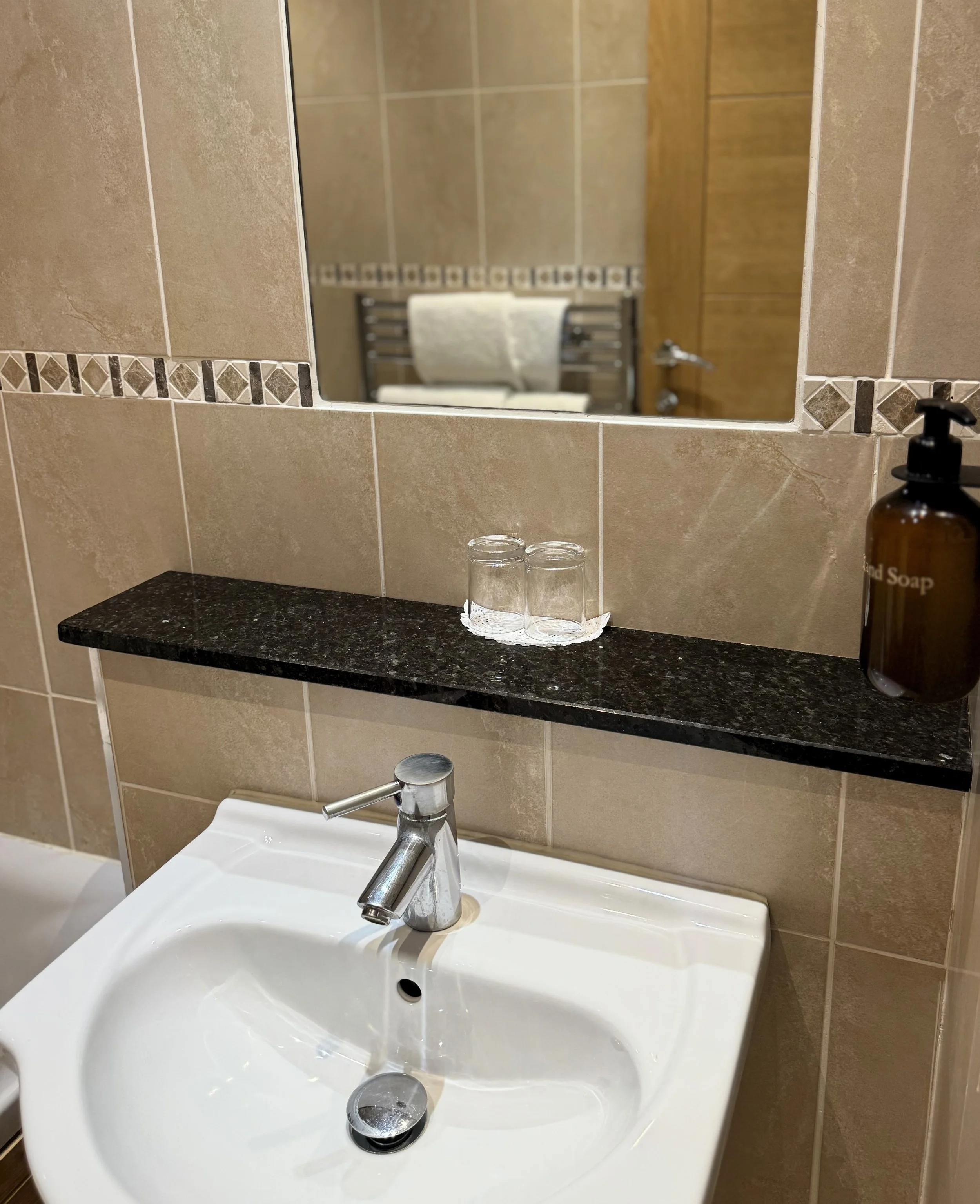 Bathroom sink with a chrome faucet, a soap dispenser on the right, and a black granite shelf mounted on beige tiled wall above the sink, holding two glasses and a soap tray, with a mirror reflecting a towel rack and wooden door.