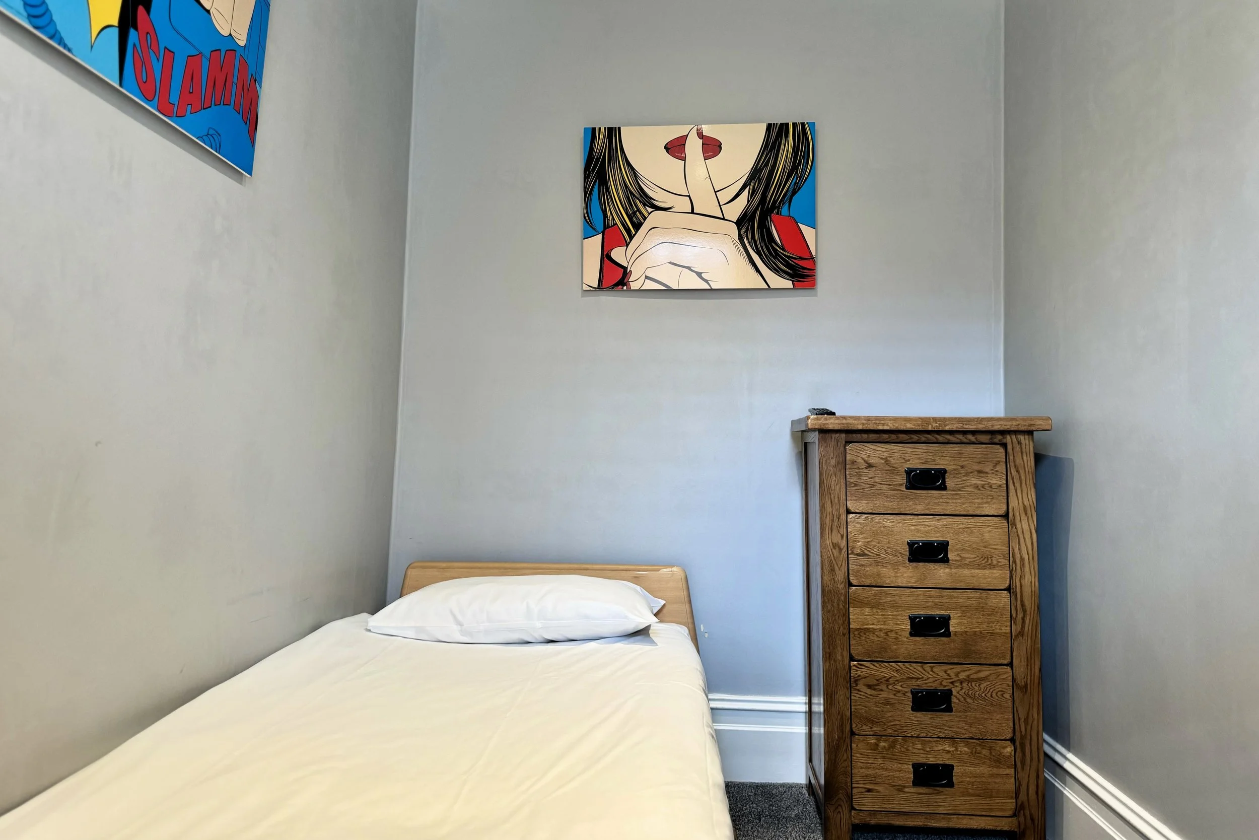 A small bedroom with a simple bed covered in white bedding, a wooden dresser, and comic-style artwork on light gray walls.