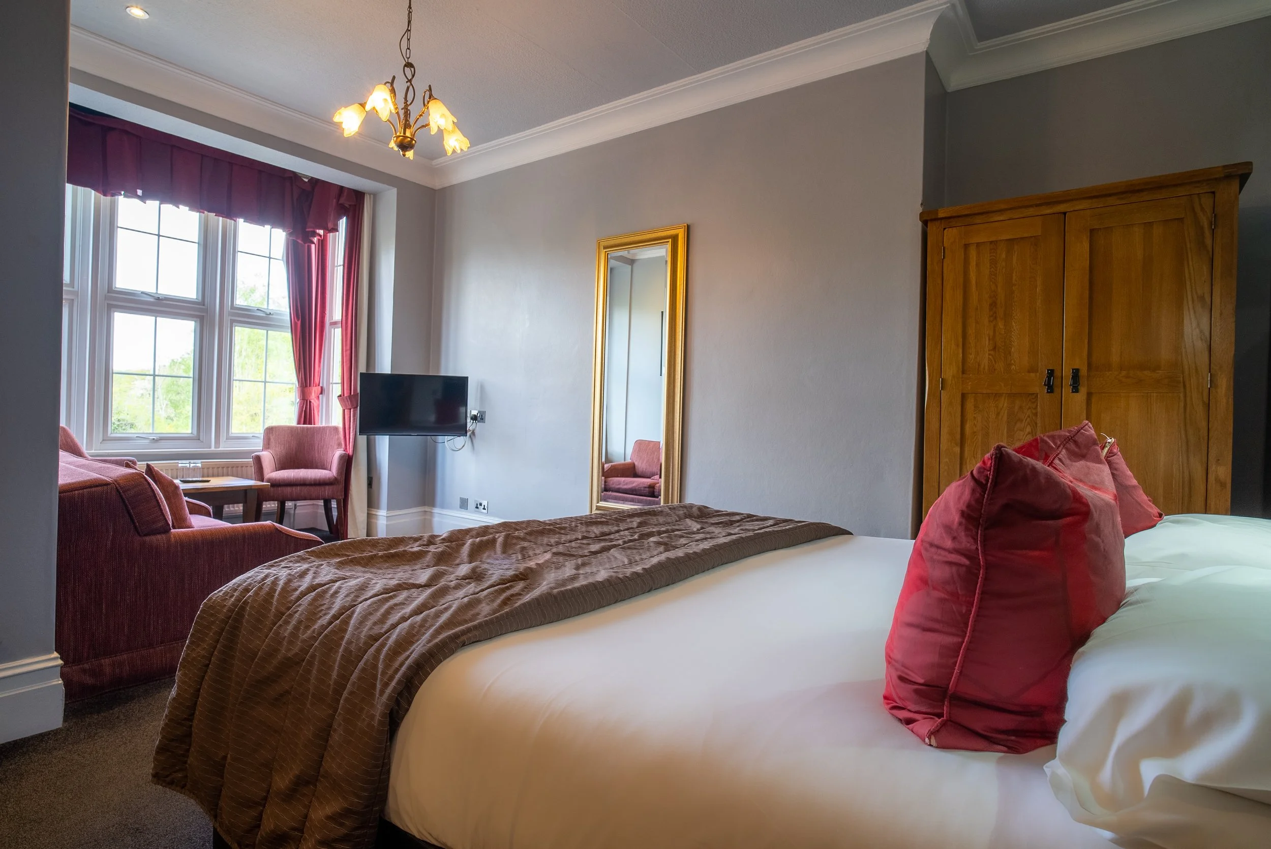 A hotel room with a large bed, pink pillows, brown bed runner, window with pink curtains, a small seating area with two pink chairs and a table, a flat-screen TV, a tall mirror with a gold frame, a wooden wardrobe, and a ceiling light fixture.