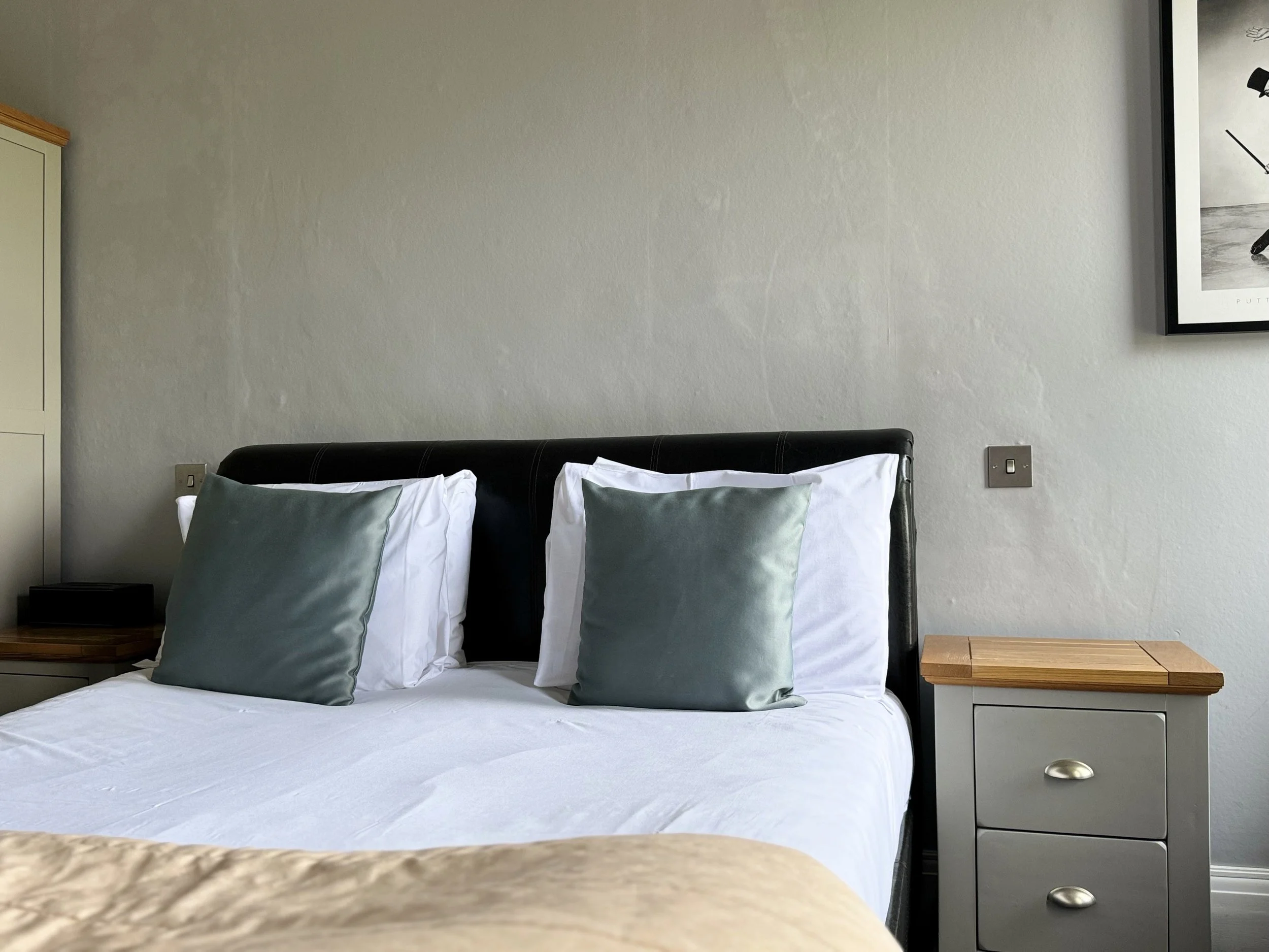 A neatly made bed with white sheets, two pillows with white pillowcases, and two decorative gray pillows, flanked by wooden nightstands, against a plain white wall with a framed black and white photograph of a person playing golf.