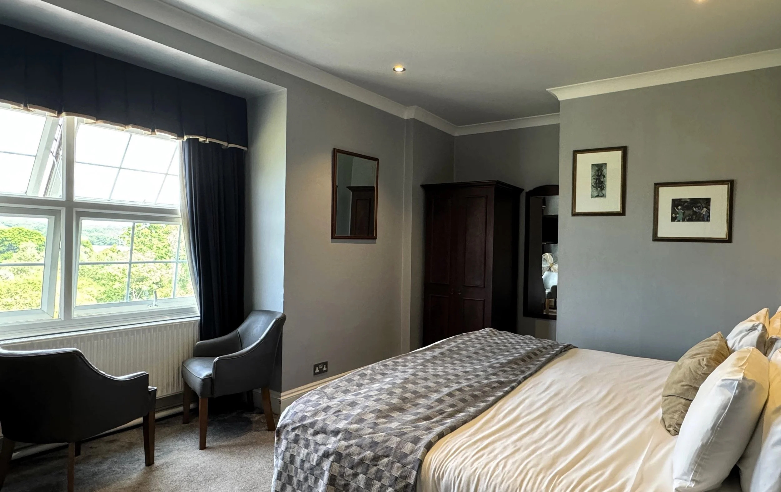 A bedroom with a bed, two black armchairs by a large window with curtains, and framed pictures on the wall.