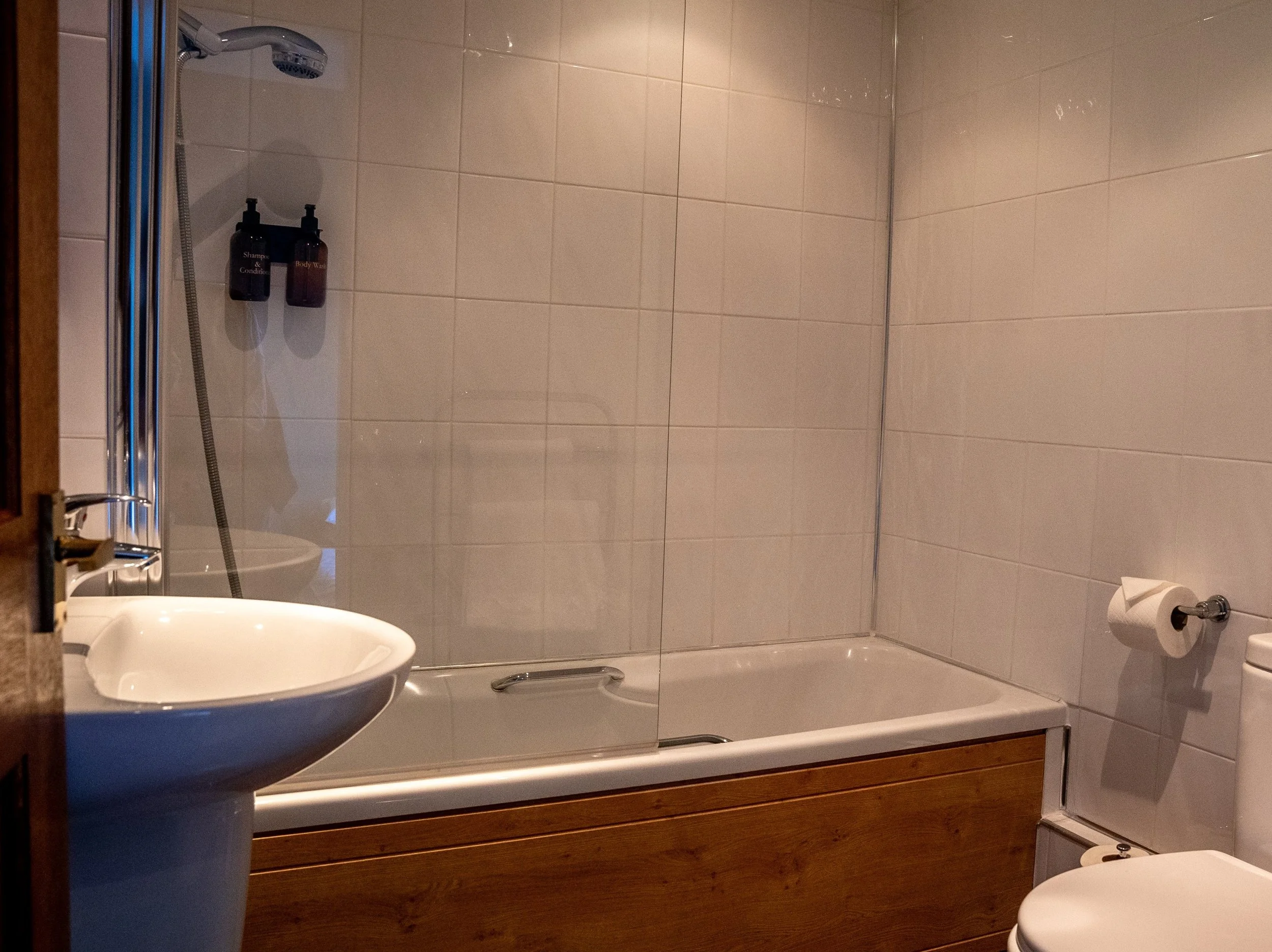 Bathroom with a bathtub, shower, sink, and toilet, featuring beige tiled walls and a wooden panel around the bathtub.