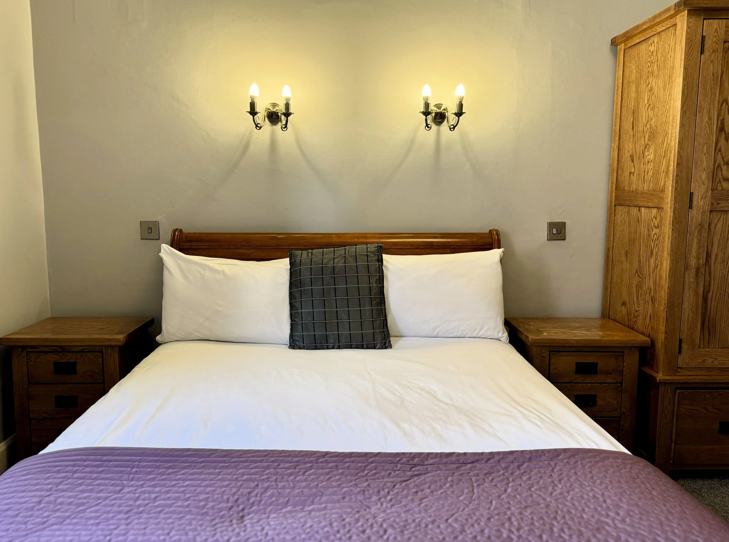 A neatly made bed with white sheets, a pinkish-purple quilt at the foot, and three pillows, one decorative pillow with a black and gray checkered pattern, against a beige wall with two wall-mounted light fixtures with five bulbs each. There are woode