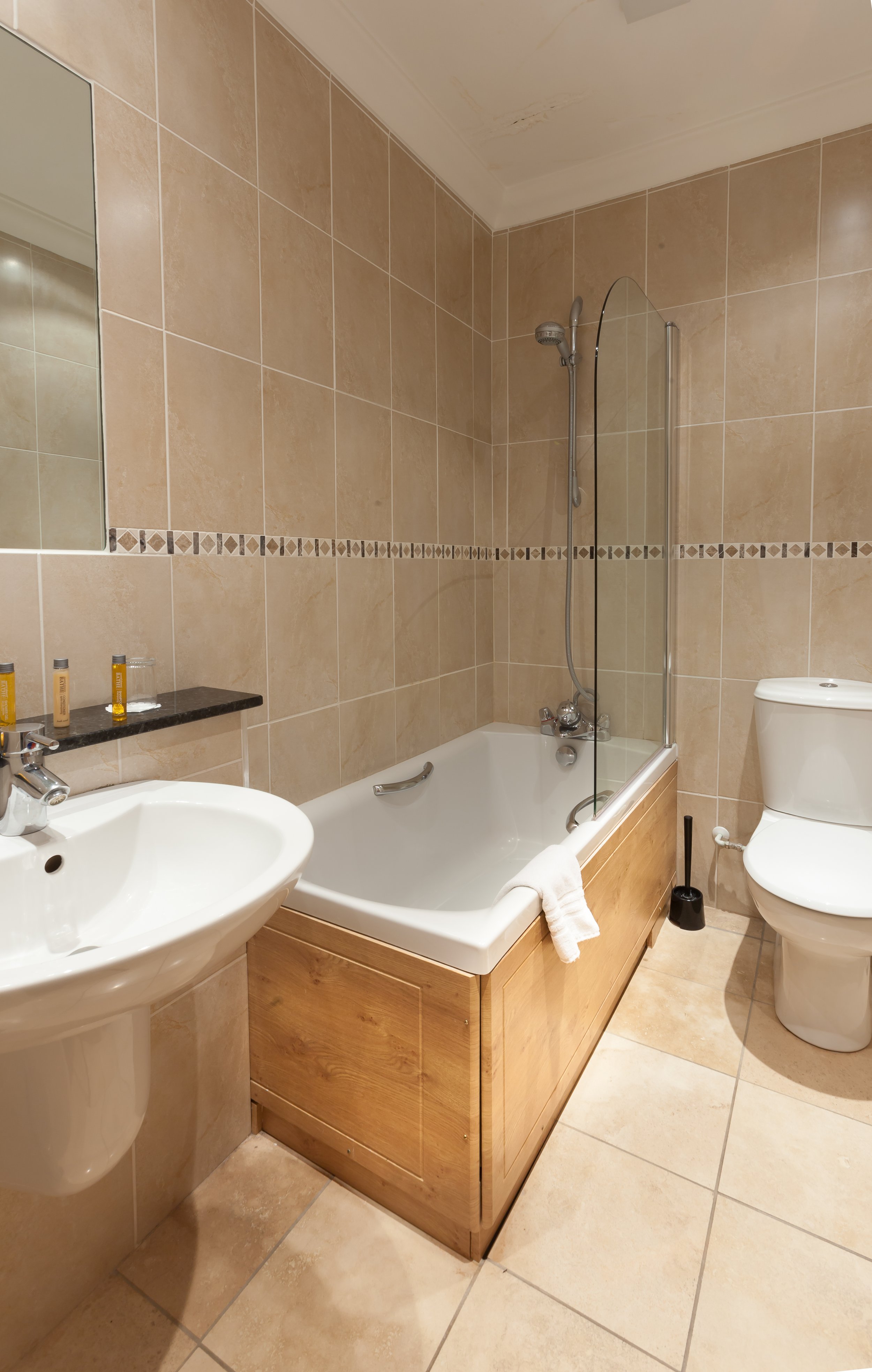 Bathroom with beige tiled walls and floor, a white bathtub with a wooden panel, a glass shower screen, a white toilet, and a white sink with a black shelf holding small toiletries.