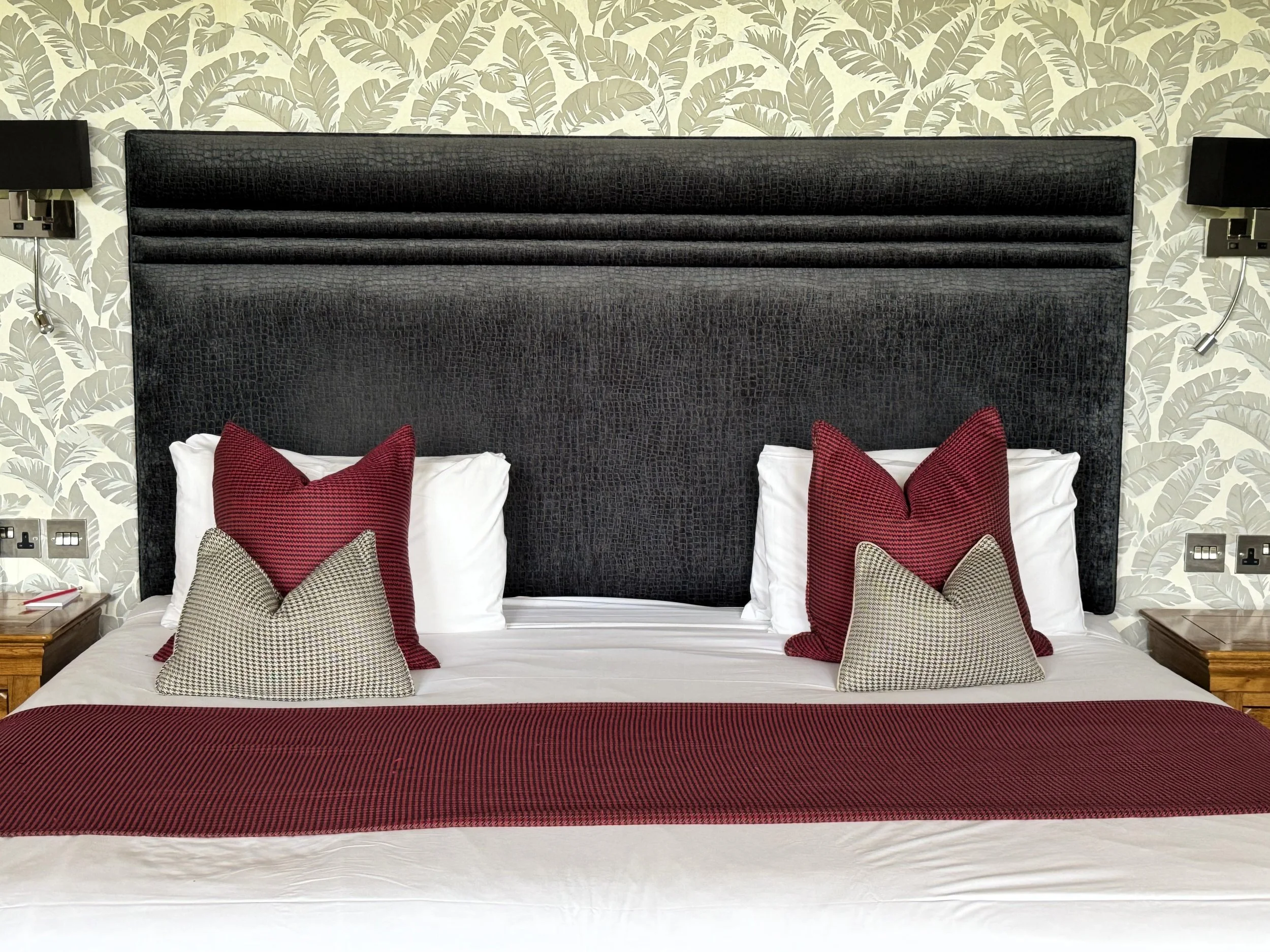 A neatly made hotel bed with a dark textured headboard, four decorative pillows in red and beige, a red runner, and matching pillows on a white bedspread. Two nightstands with lamps and wall outlets are visible on either side.