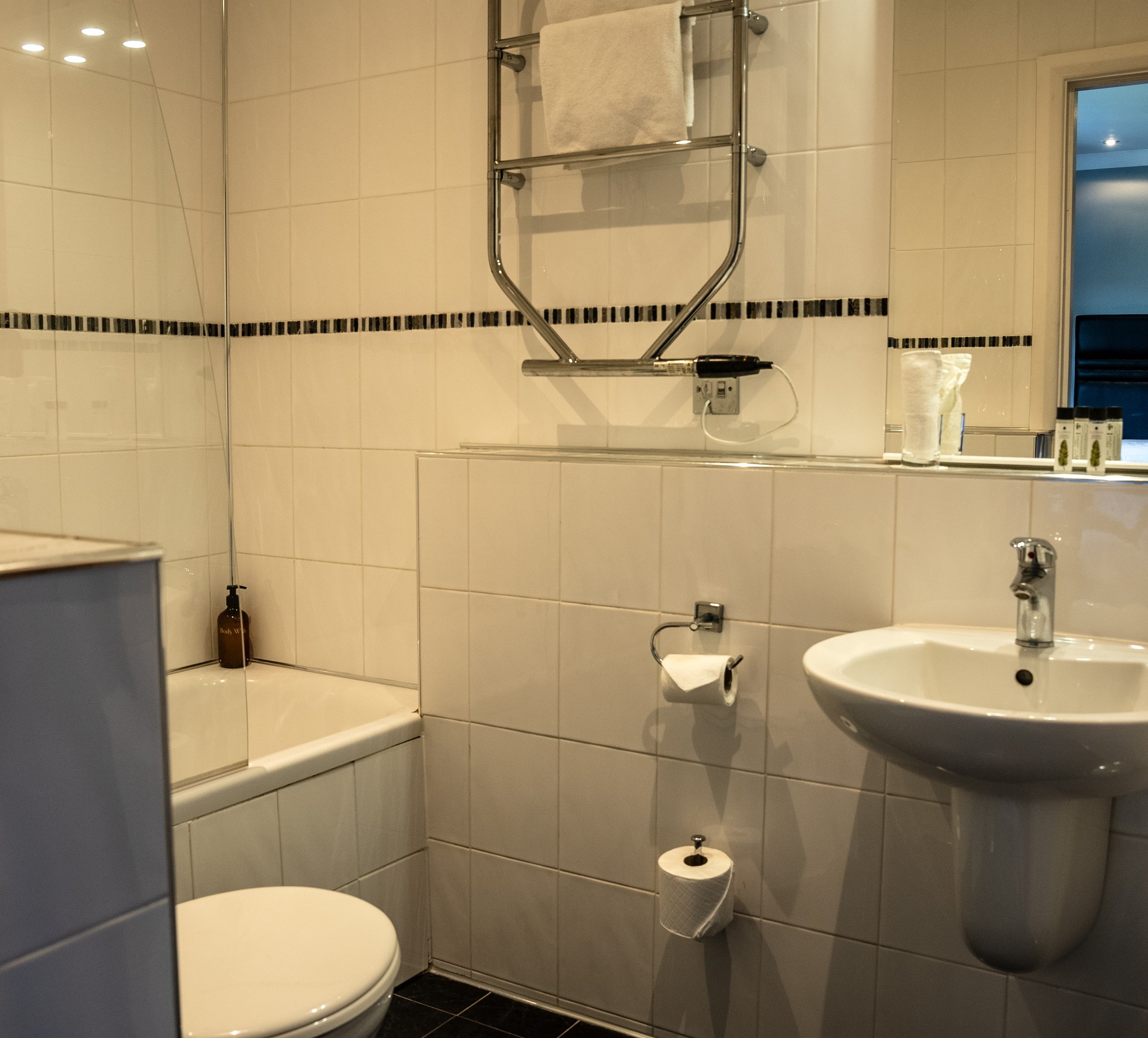 Small bathroom with a toilet, sink, mirror, and a shower area with a glass partition. Towel hanging on a wall-mounted towel rack, and there are toiletries and a soap dispenser visible.