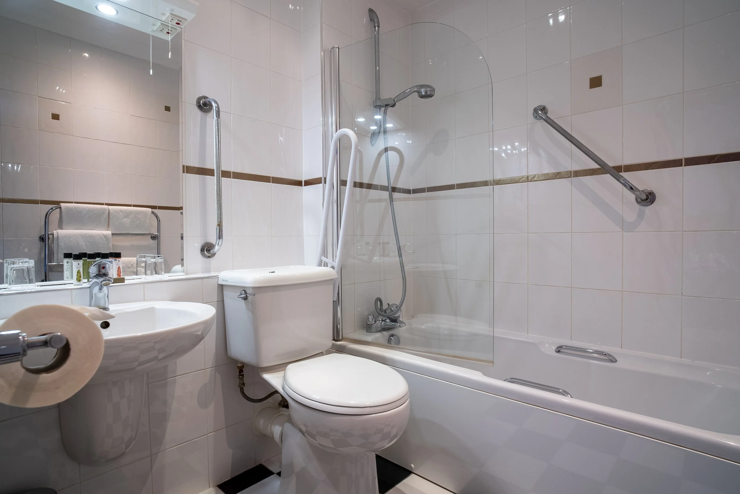 Bathroom with white tiled walls, a toilet, a small sink, a bathtub with a glass shower screen, and grab bars for assistance.