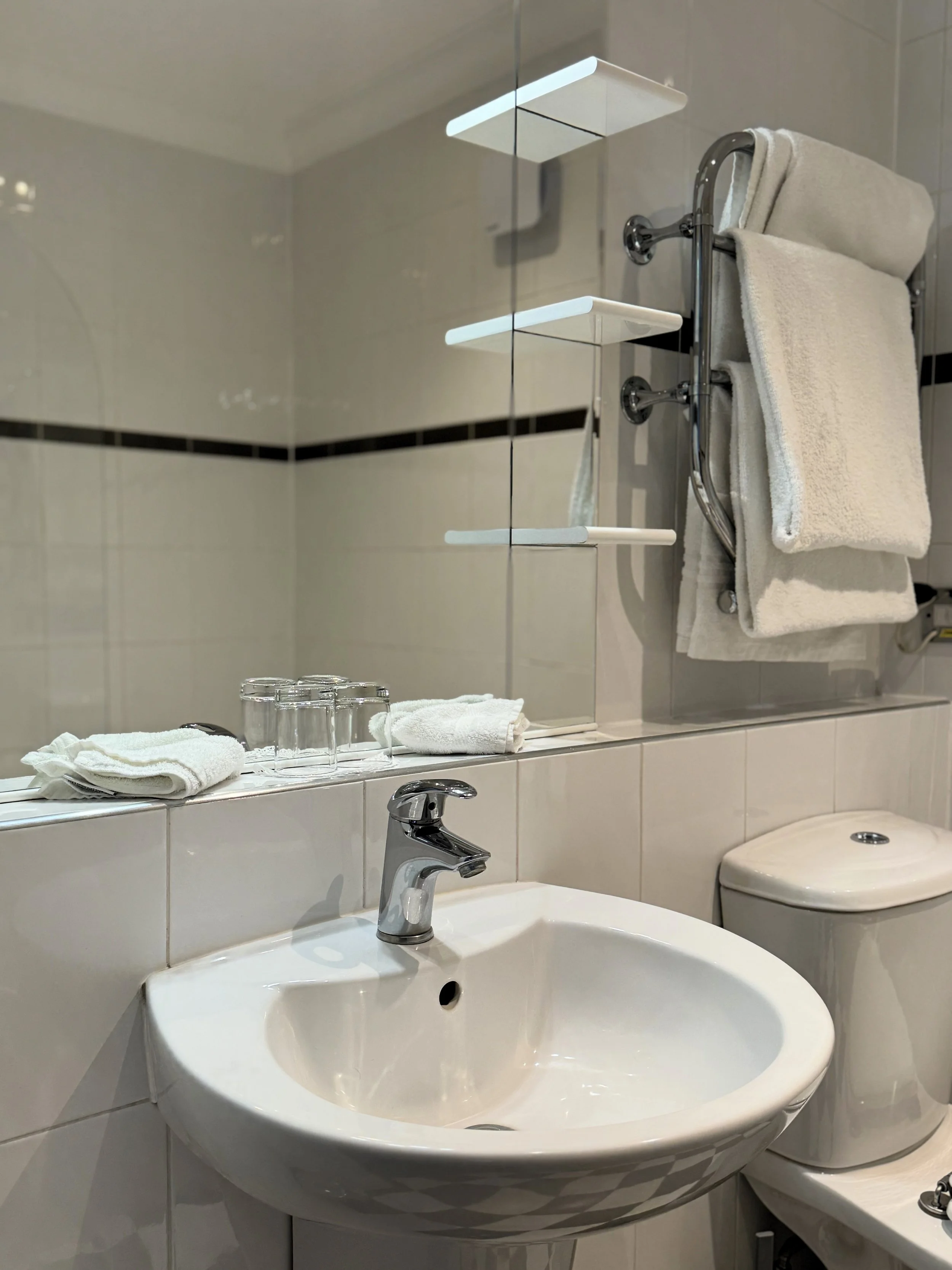 A bathroom with a white sink, mirror, toilet, and a wall-mounted towel rack holding folded towels. Glasses and small towels are placed on the ledge above the sink.
