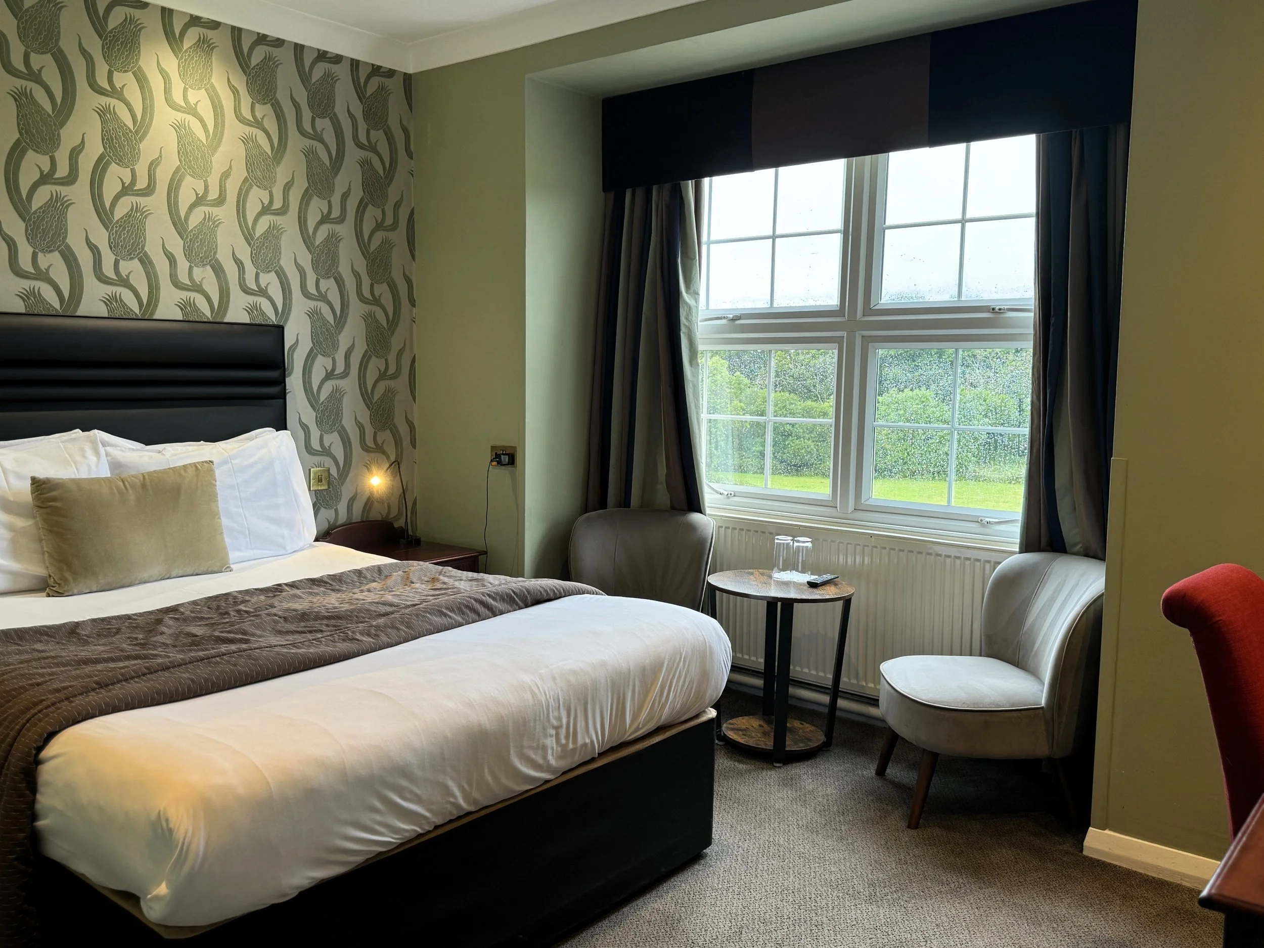 Hotel room with a bed, a chair, a window with curtains, and a small table with glasses and a remote.