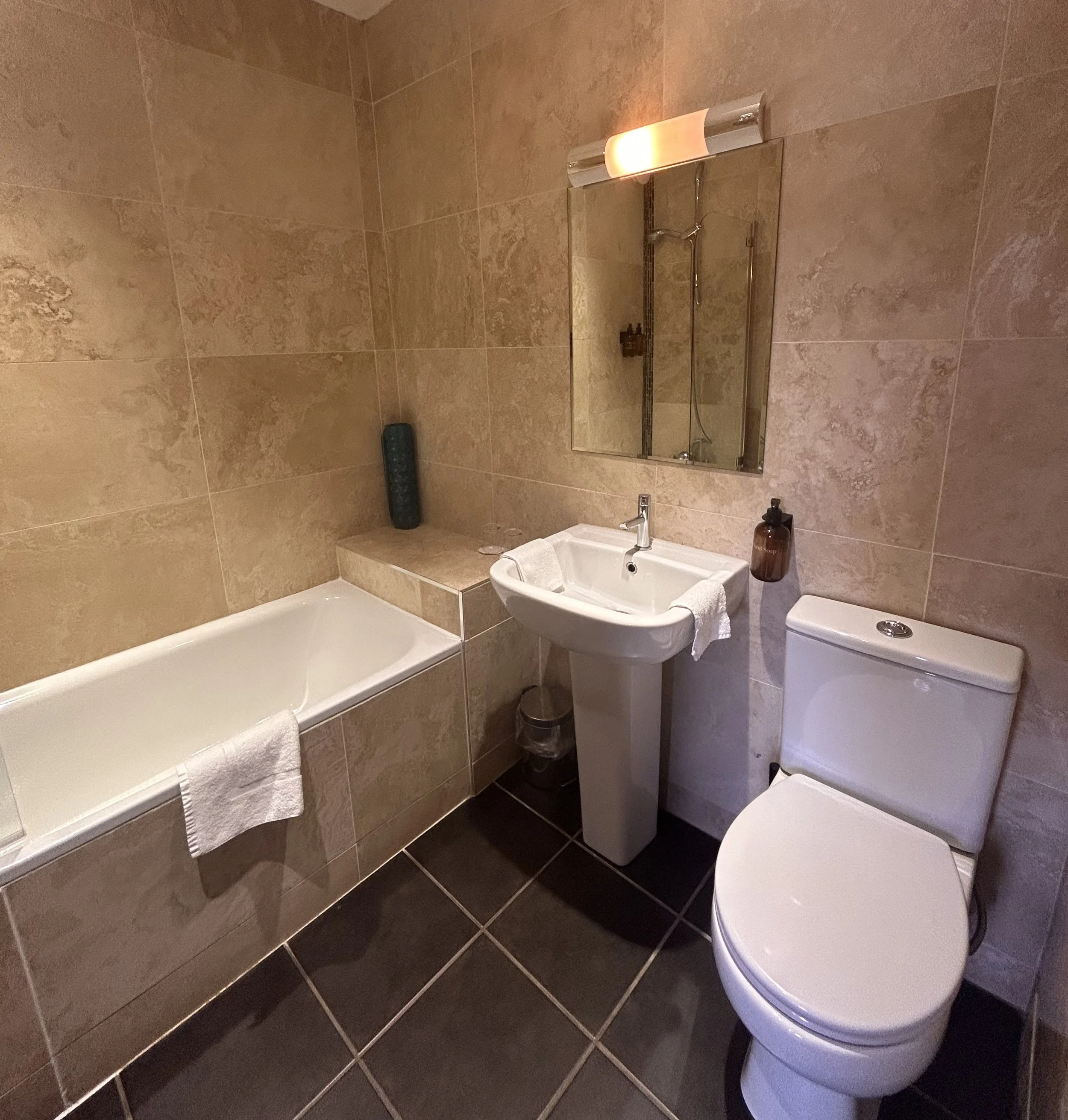A modern bathroom with a bathtub, pedestal sink, and toilet, featuring beige tiled walls and dark tiled floor, with a mirror above the sink.