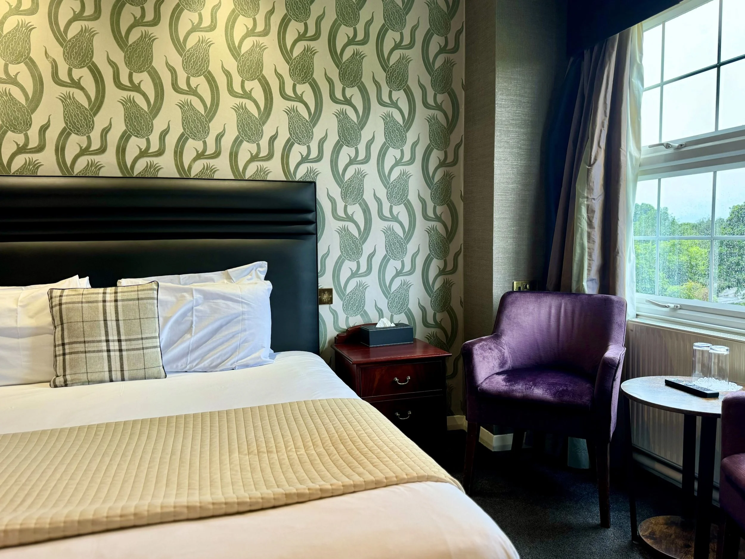 A hotel room with a bed, a purple armchair, a round side table with two glasses and a remote, and a window with green trees outside. The wall behind the bed has a green and beige botanical patterned wallpaper.