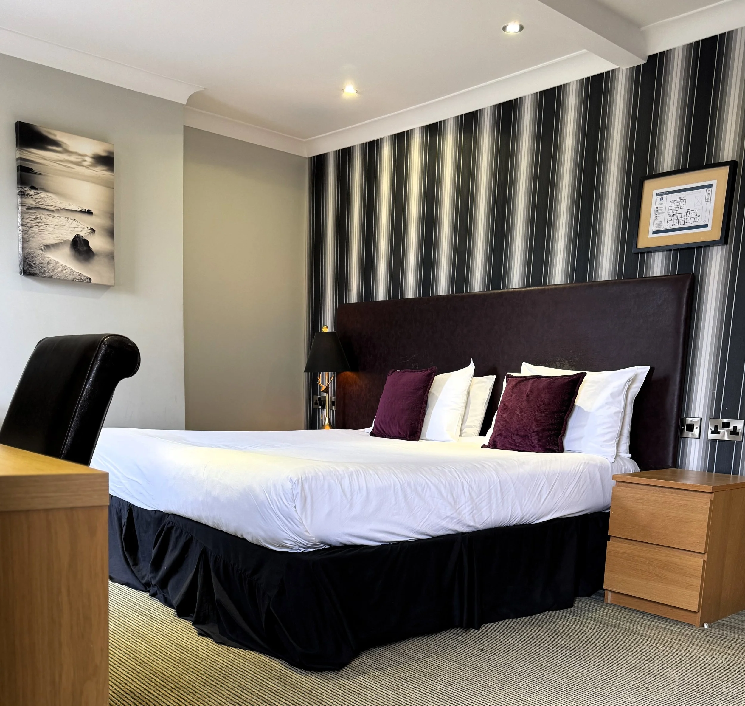 A modern hotel room with a bed that has white sheets, purple accent pillows, and a tall purple headboard. There is a black table lamp on the right side of the bed, wooden nightstands, a black leather chair, a striped wall behind the bed, and a framed