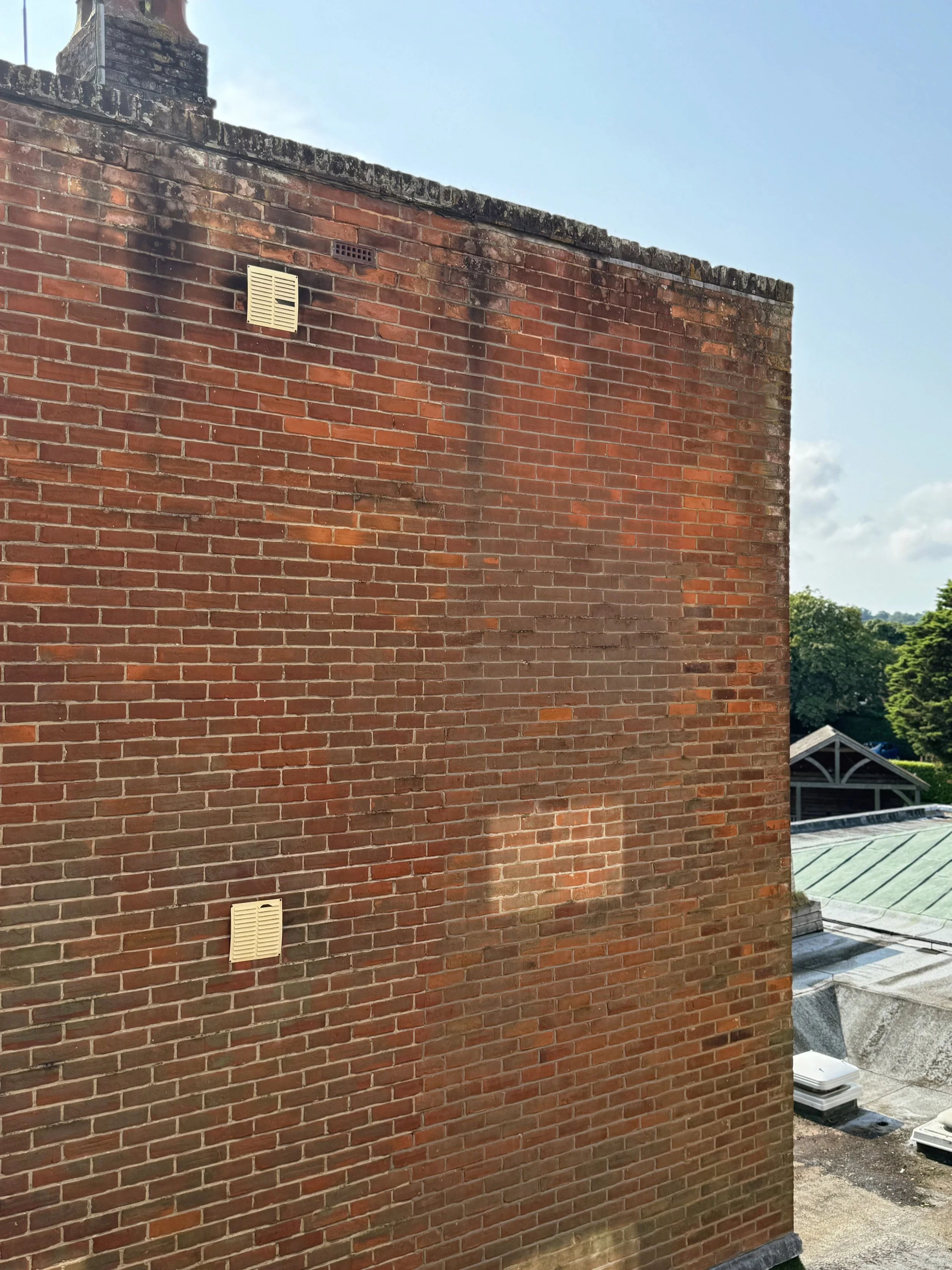 Close-up of a brick building wall with two beige air vents, black soot stains, and a chimney at the top, with trees and a cloudy sky in the background.